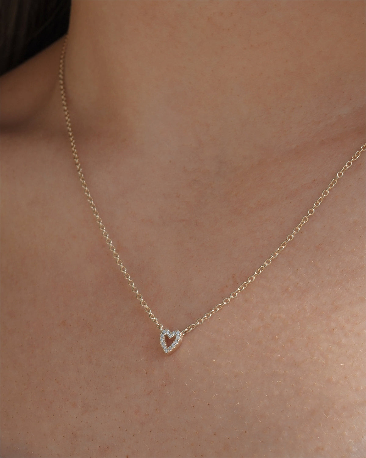 A delicate gold chain necklace with a small open heart pendant encrusted with lab grown diamonds, shown worn on a person's neck.