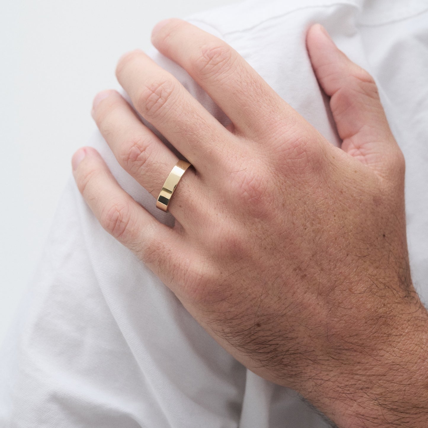 Man's hand wearing a sleek gold band, showcasing minimalist high-end jewellery, with a white shirt in the background.