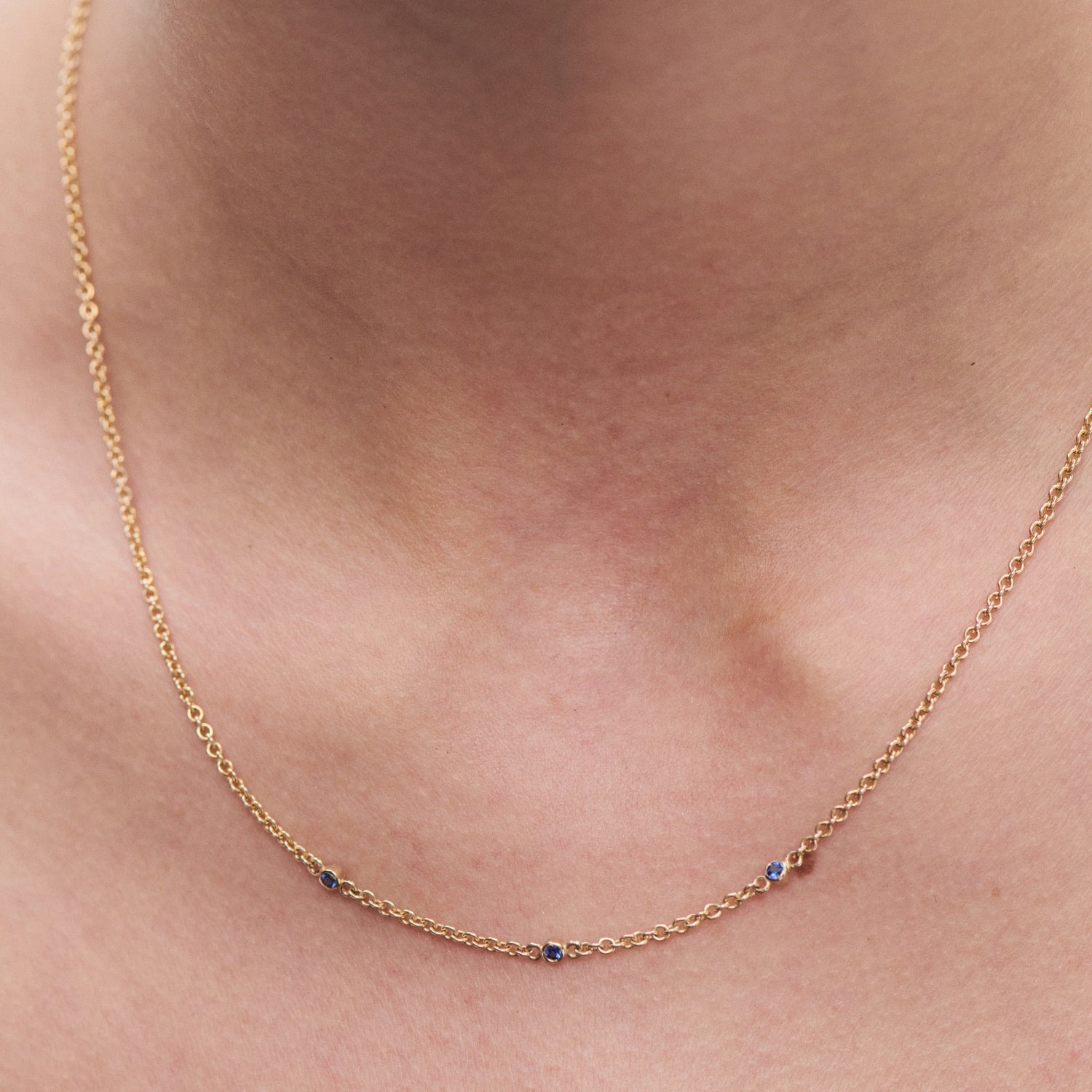 Delicate gold necklace with small blue gemstones, elegantly draped on a person's neck. Luxury jewellery, minimalist design.