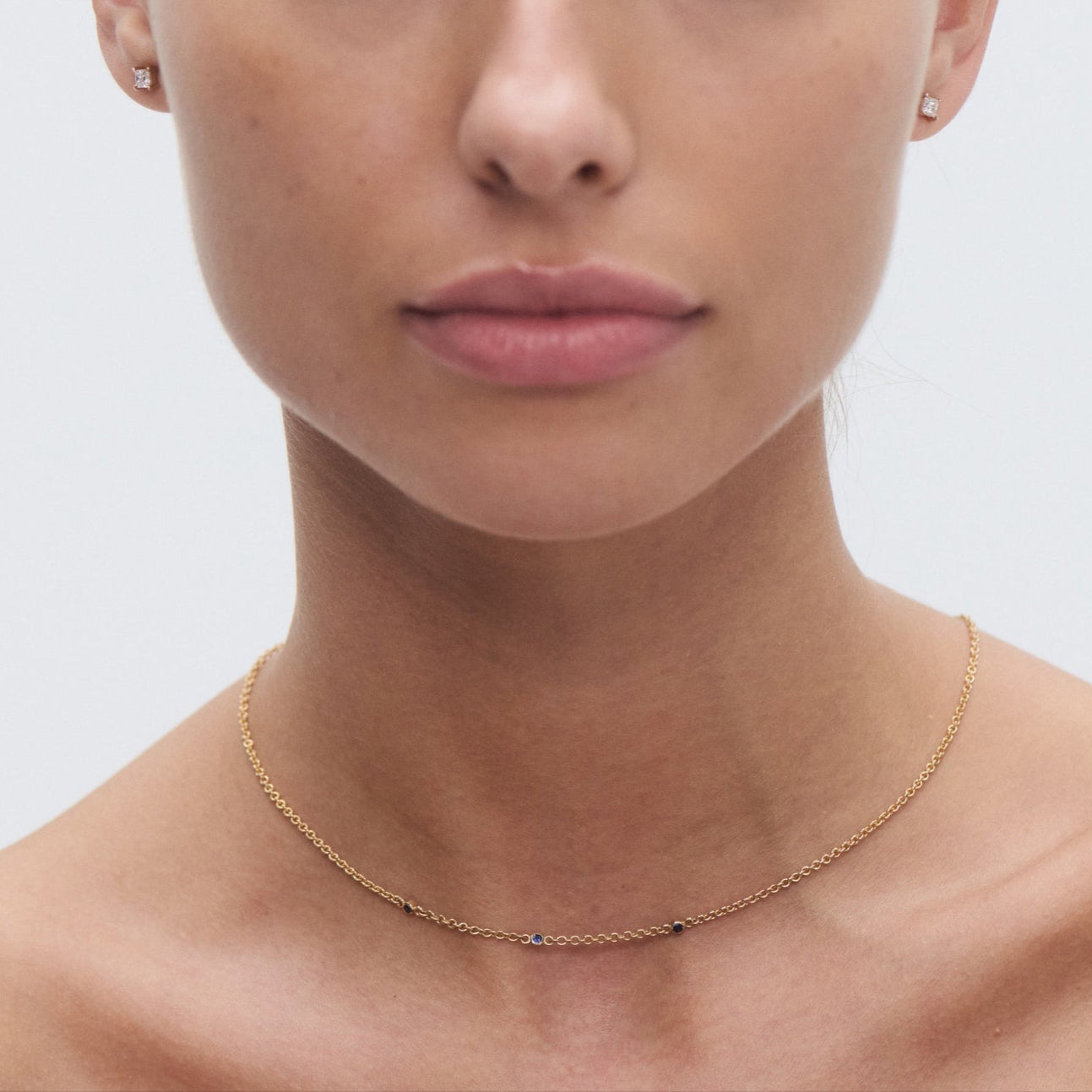 Woman wearing a delicate gold necklace with small gemstones and diamond stud earrings, highlighting luxury jewellery. Light background.