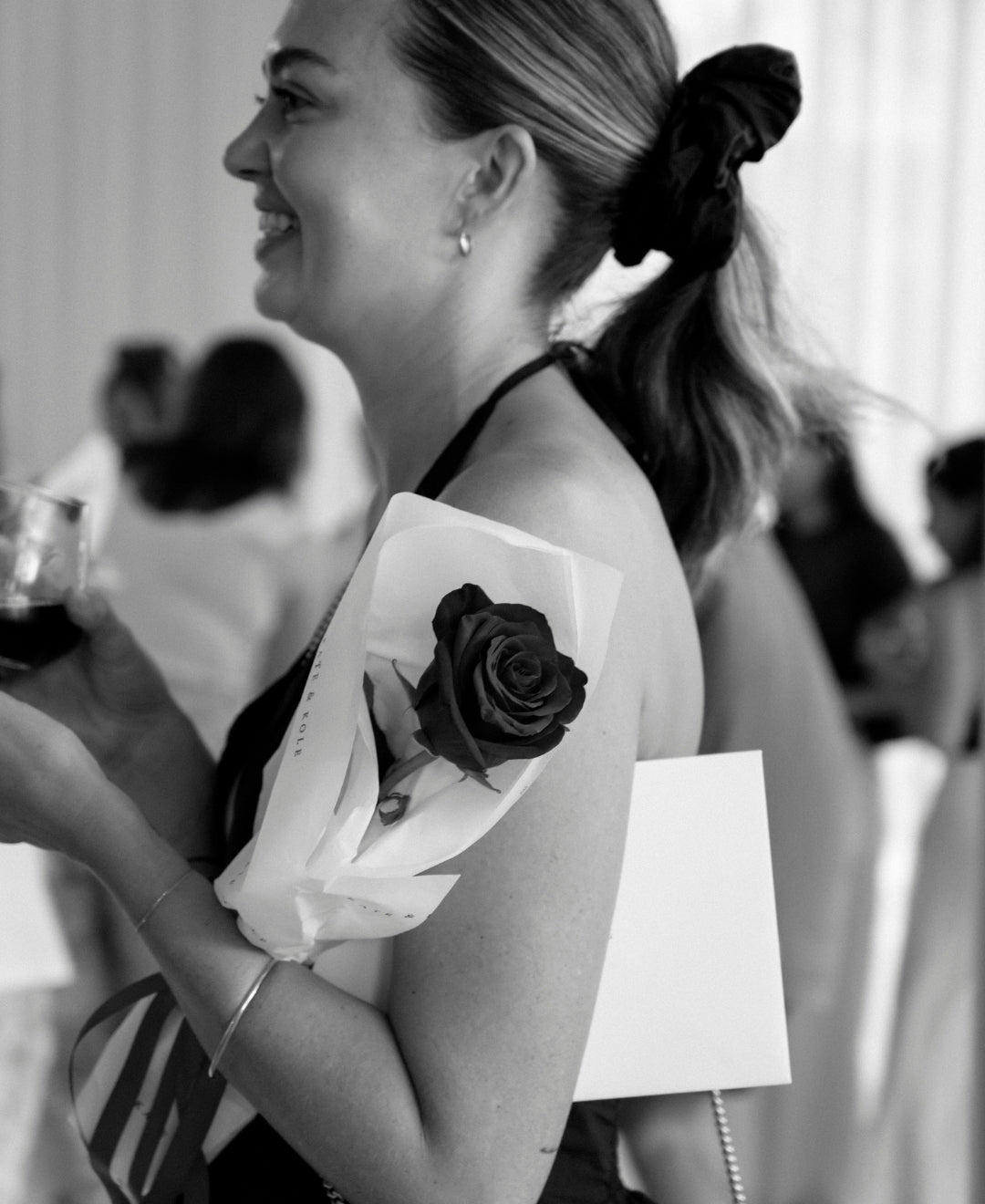 Woman smiling, wearing a black dress, holding a glass of wine and a wrapped rose. Hair tied with a scrunchie, jewellery visible. Elegant setting.