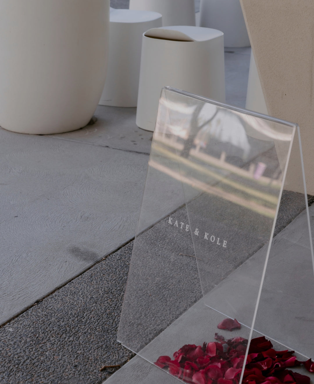 Clear sign with "KATE & KOLE" in white text on concrete, surrounded by stylish white stools and scattered red rose petals.