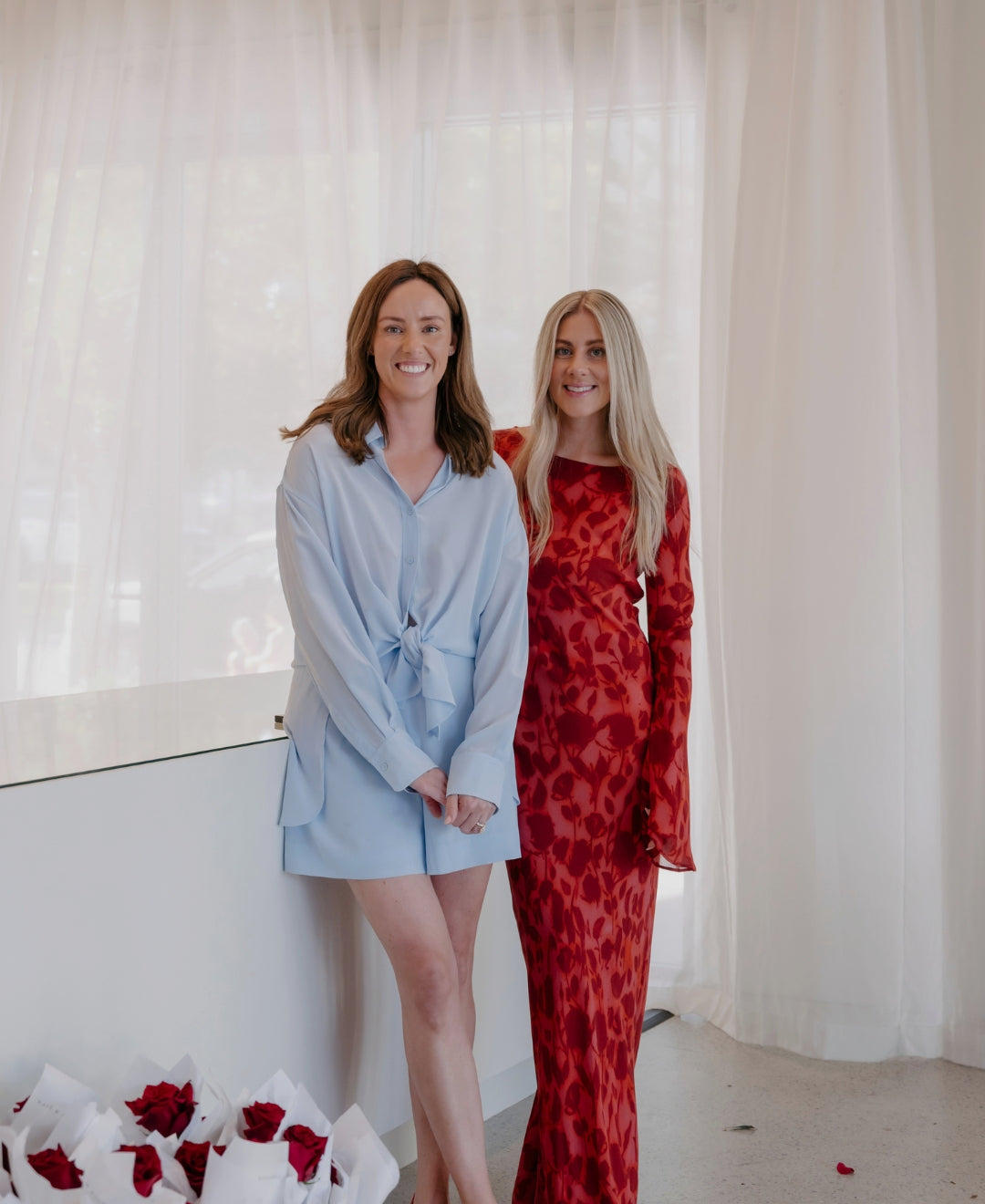 Two women stand in a chic setting; one in a light blue dress, the other in a patterned red dress, near a table with white bouquets and red roses.