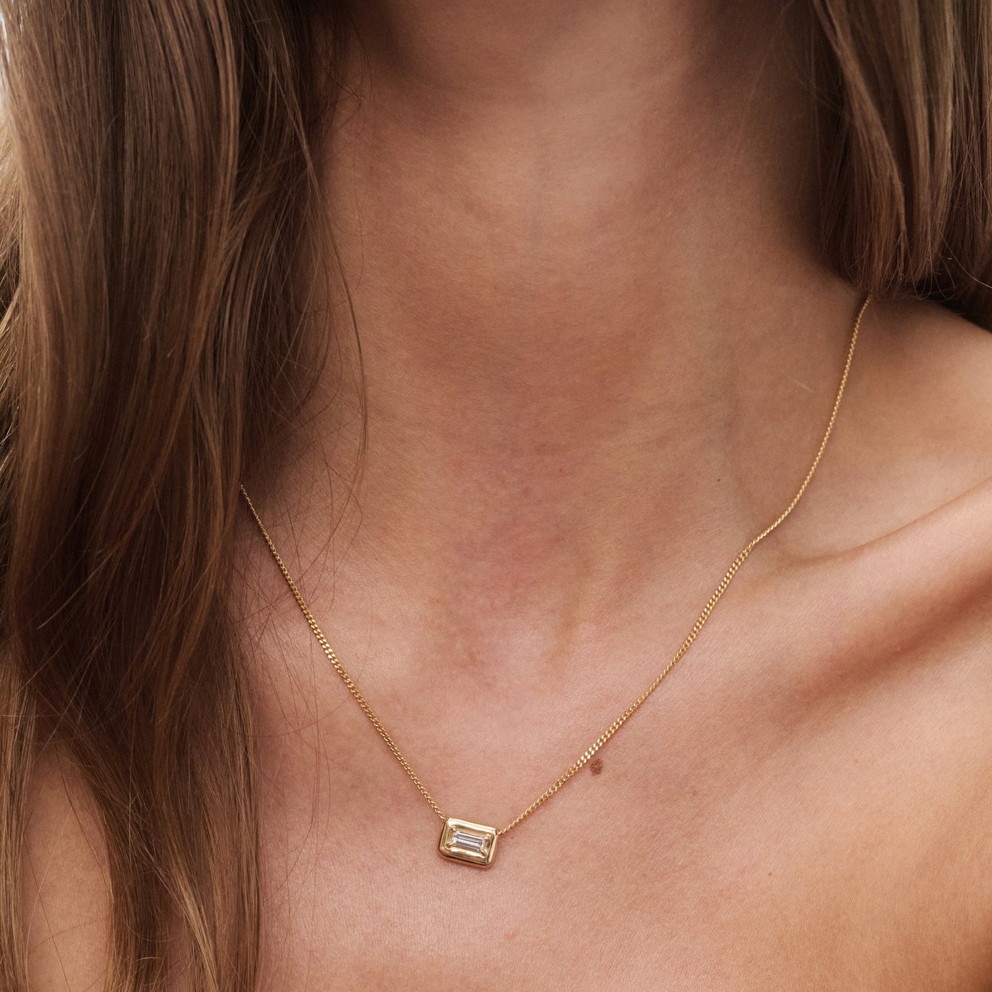 Woman wearing a gold necklace with an emerald-cut pendant. The jewellery rests elegantly on her collarbone, showcasing minimalist luxury.