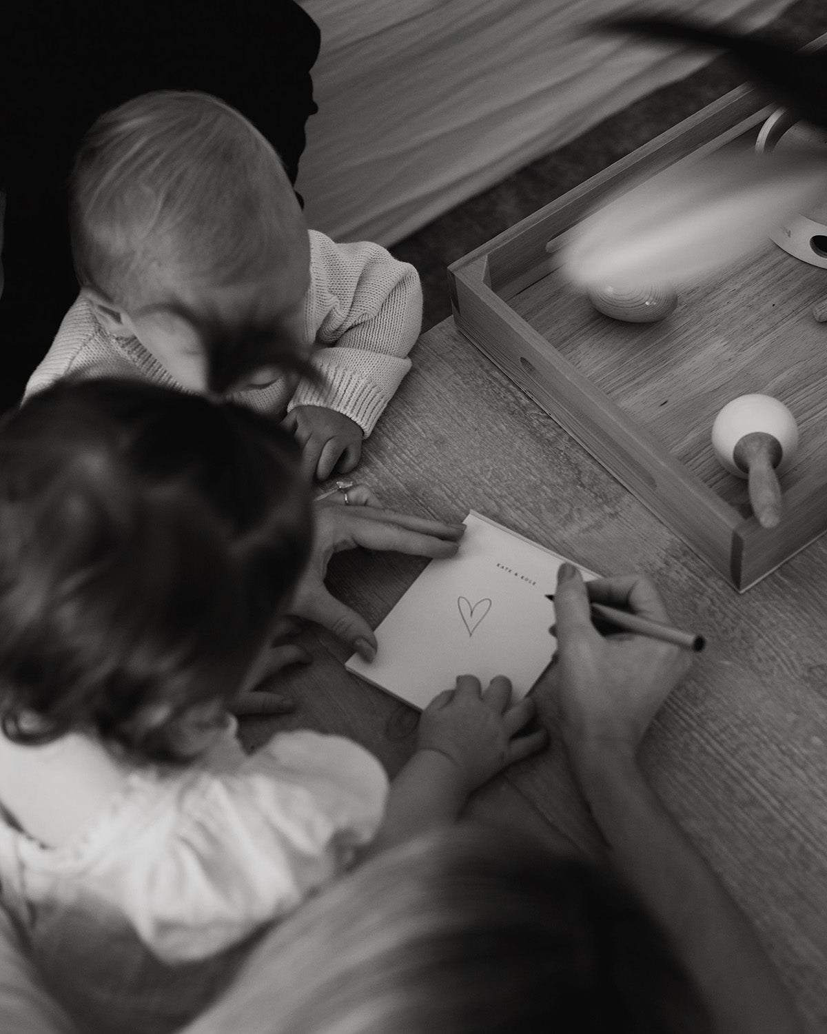 A child draws a heart on paper with help from an adult, while another child watches. A wooden tray with toys is nearby on a table.