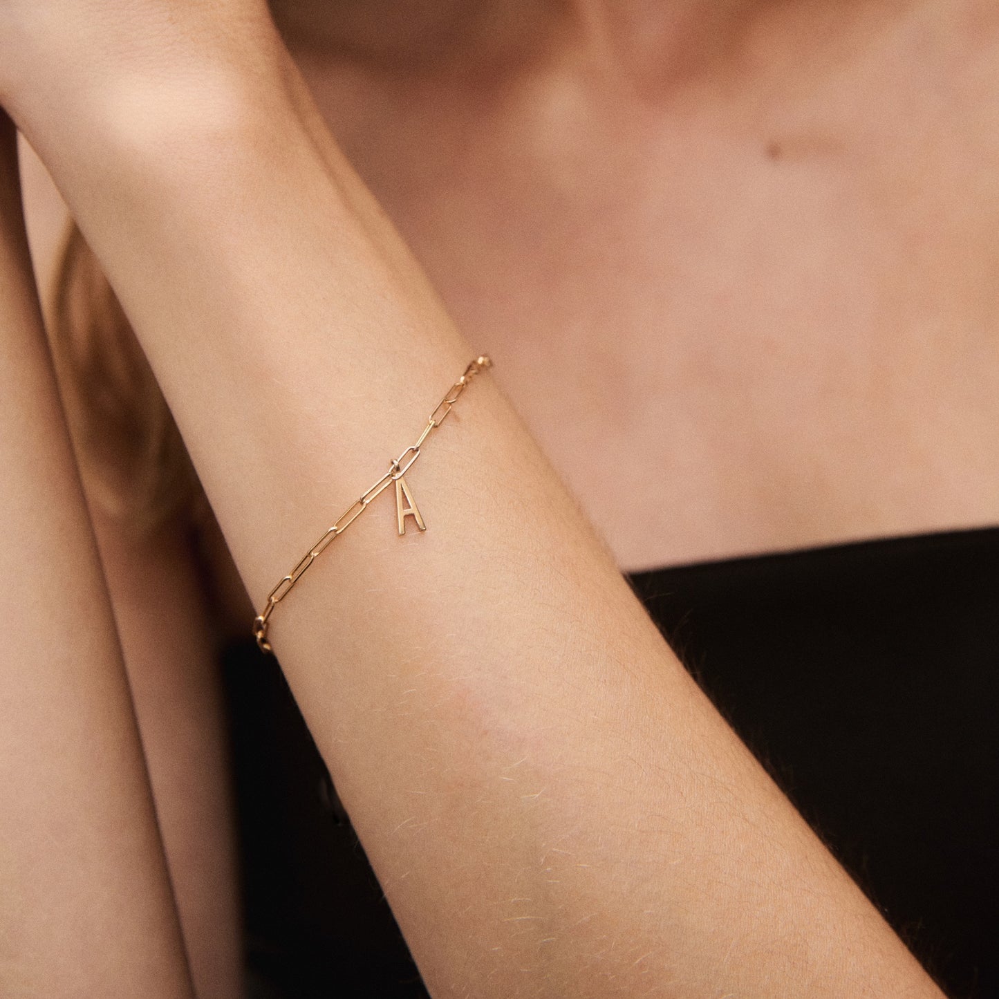Woman's arm adorned with a delicate gold bracelet featuring an "A" charm, against a black strapless top background. Elegant and luxurious jewellery.