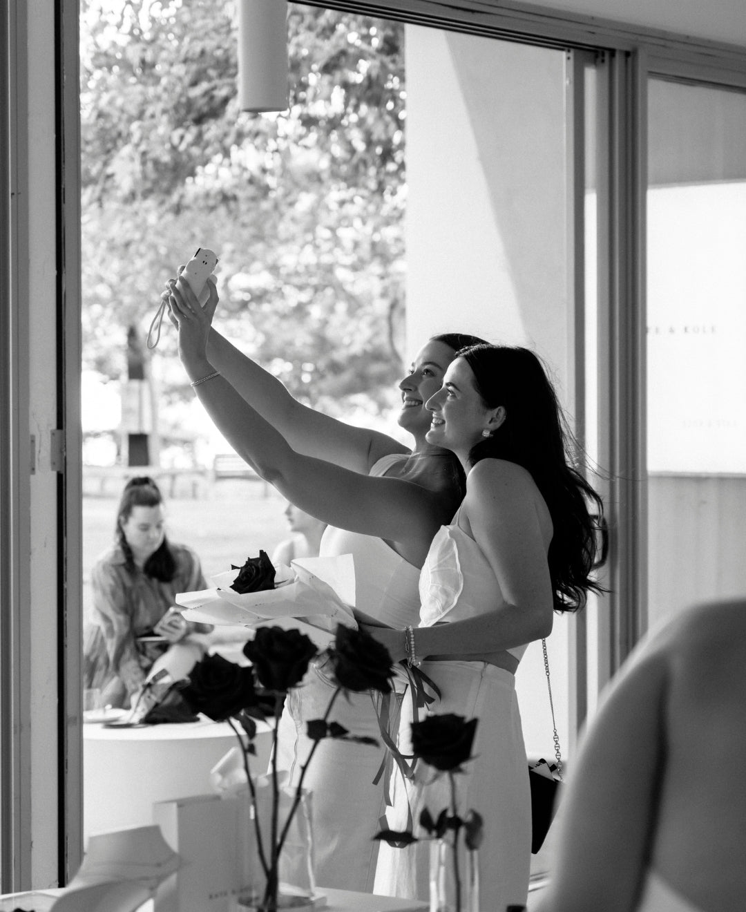 Two women in elegant outfits take a selfie in a stylish room with black roses and a large window, embodying a luxury jewellery vibe.