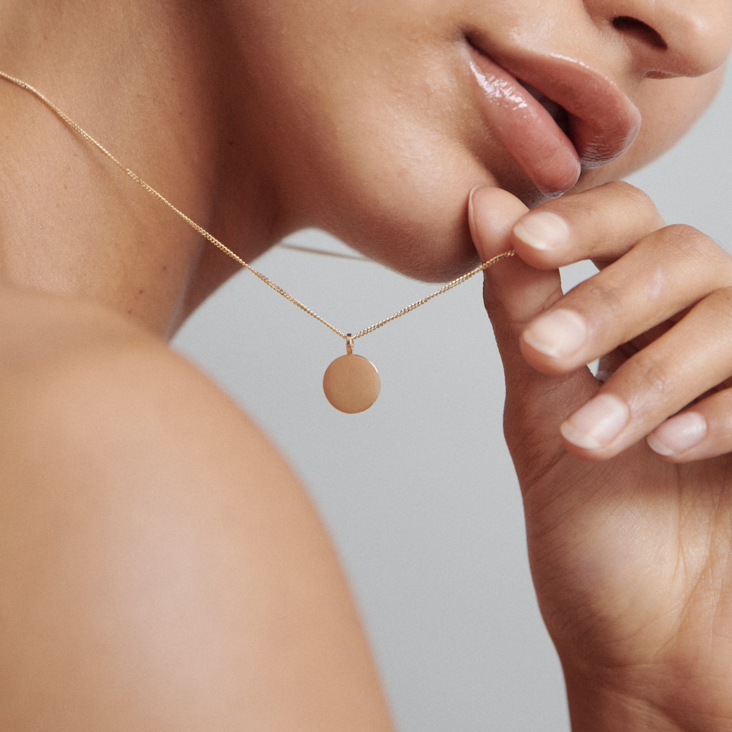 Close-up of a person wearing a gold chain with a round pendant, gently touching it. Focus on jewellery elegance and fine design.