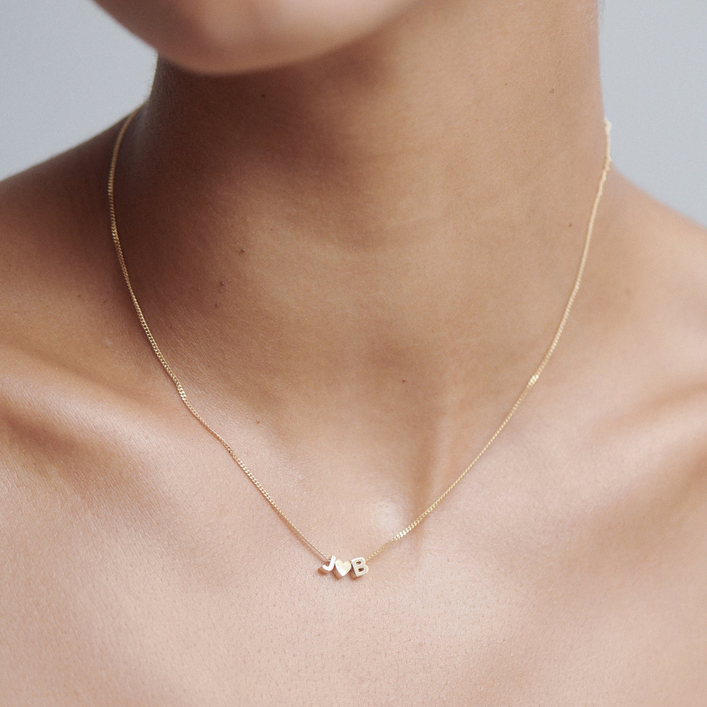 Close-up of a gold necklace with small heart and 'B' charms, worn on a person's neck. Minimalist luxury jewellery design.[text overlay: 45cm Necklace]