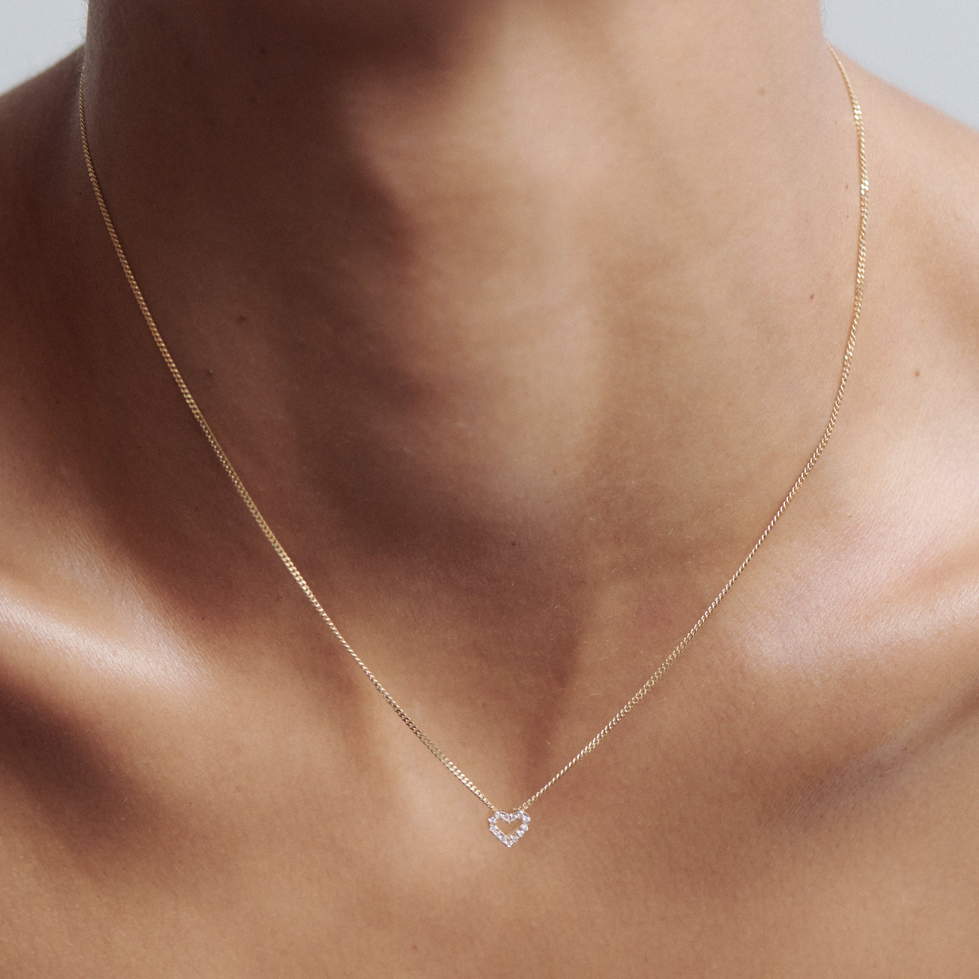Delicate gold chain necklace with a small diamond heart pendant, worn on a woman's neck. Minimalist luxury jewellery.