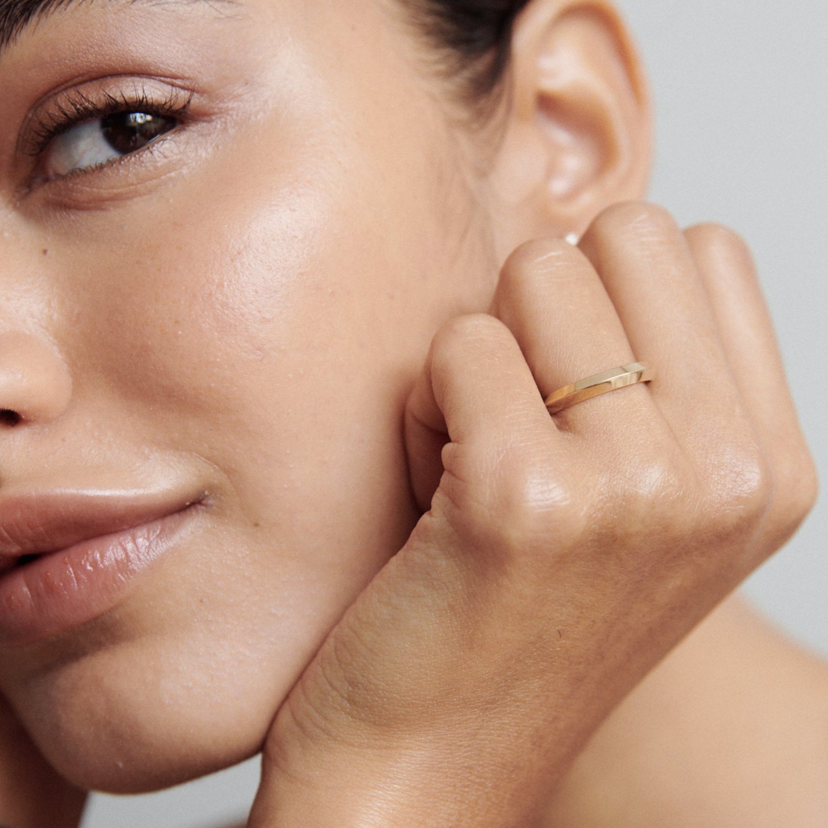 Woman's hand gracefully resting by face, showcasing a sleek gold ring. Elegant high-end jewellery captured in soft light.