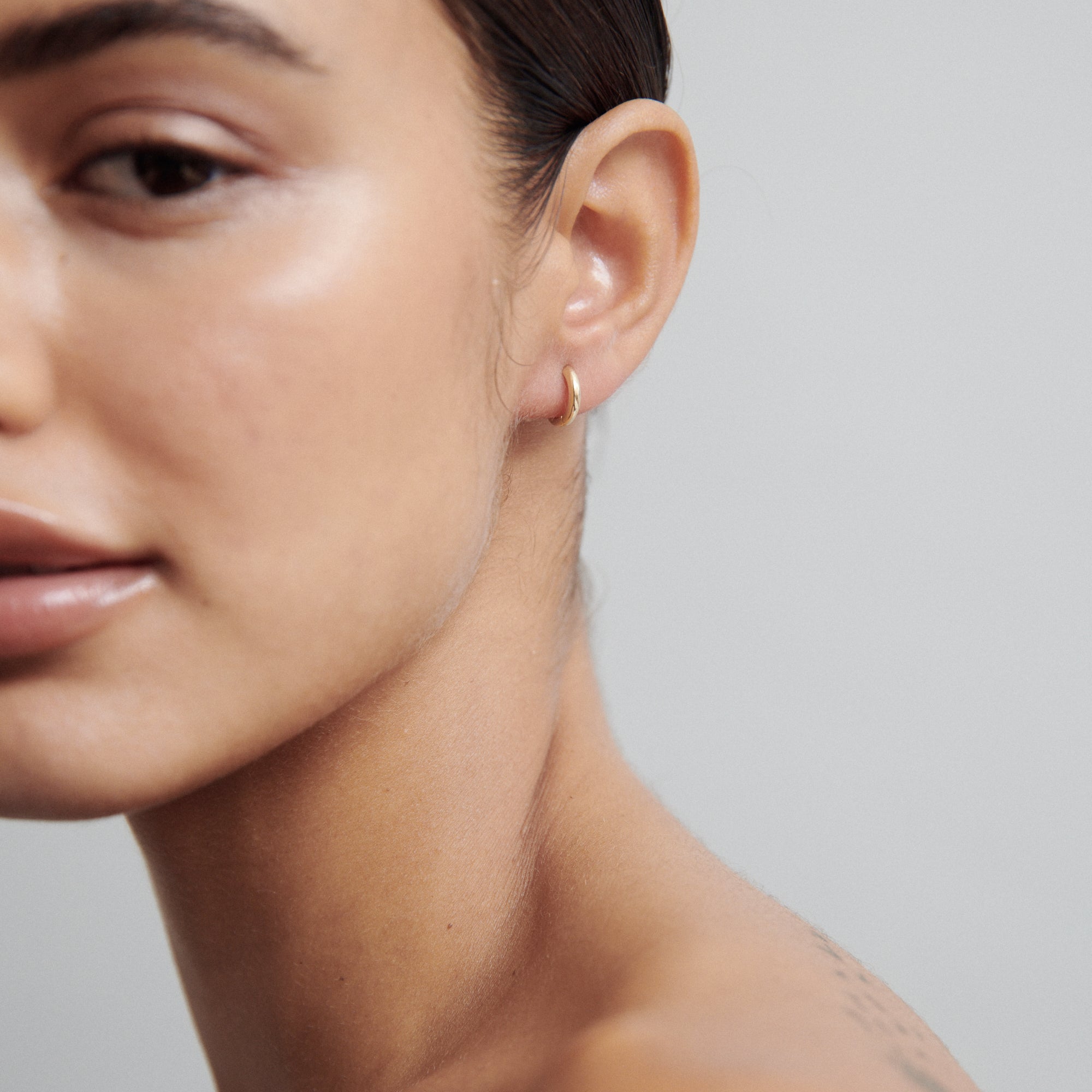 Close-up of a woman wearing a small gold hoop earring. Her skin is smooth, and the background is a soft, neutral grey.