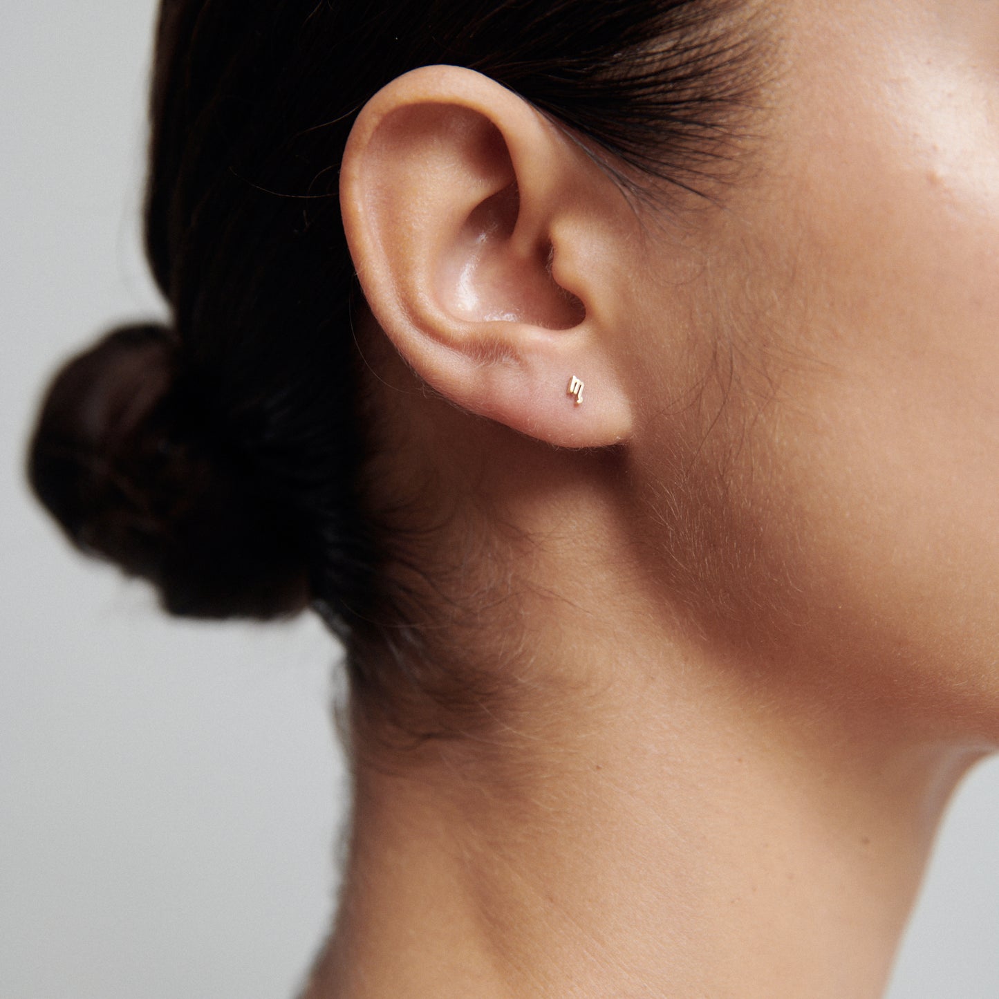 Close-up of a woman's ear wearing a small, elegant stud earring. Hair is tied in a bun, showcasing a minimalist jewellery style.