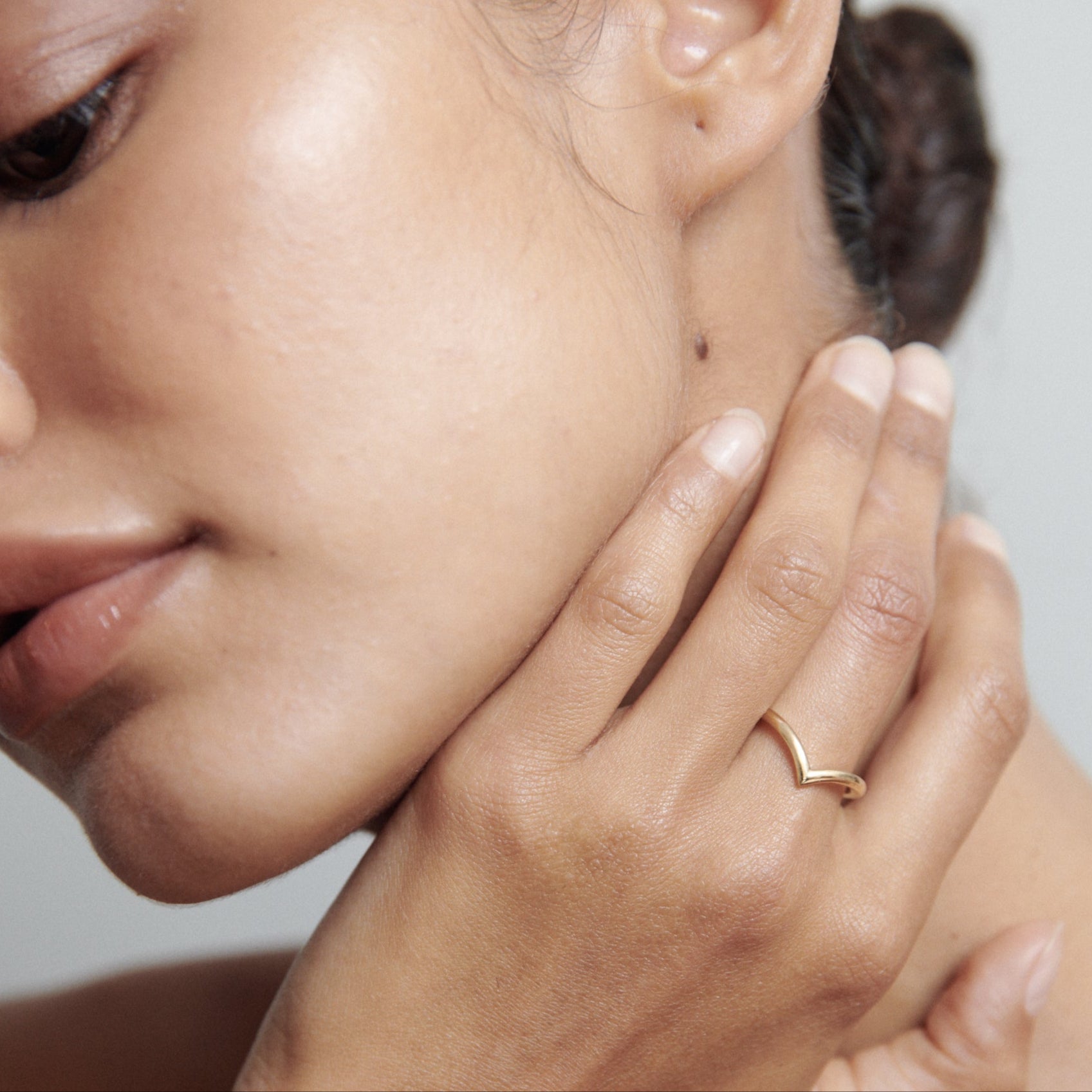 Close-up of a woman's hand gently touching her face, showcasing a minimalist gold ring. The background is a soft grey.