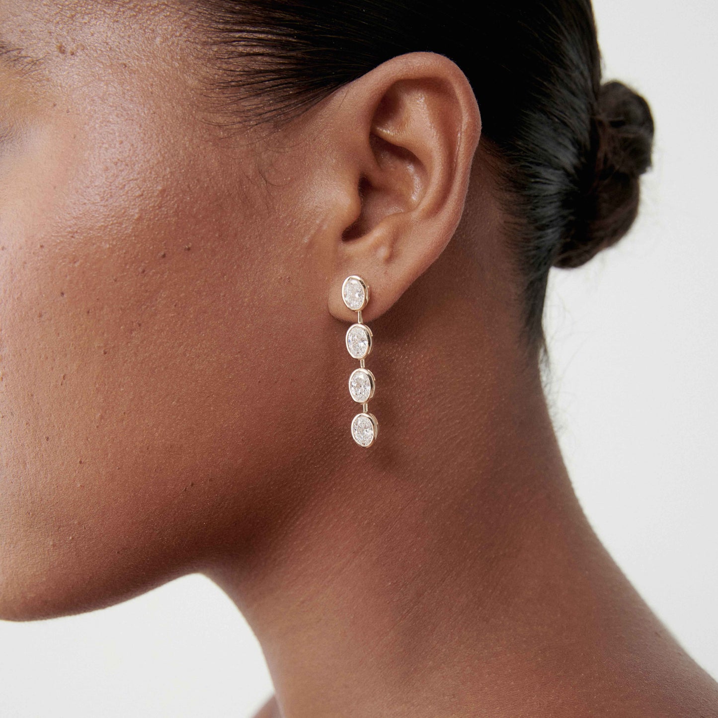 Close-up of a woman's ear wearing elegant drop earrings with four oval diamonds, set in gold. Hair is tied back for a sophisticated look.