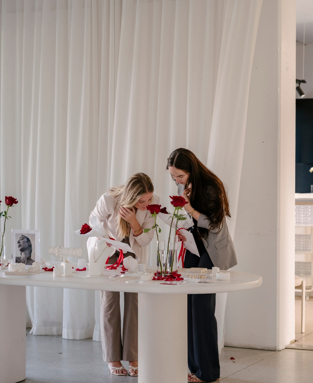 Two women in elegant attire examine jewellery on a white table adorned with red roses in a luxury setting with white curtains.