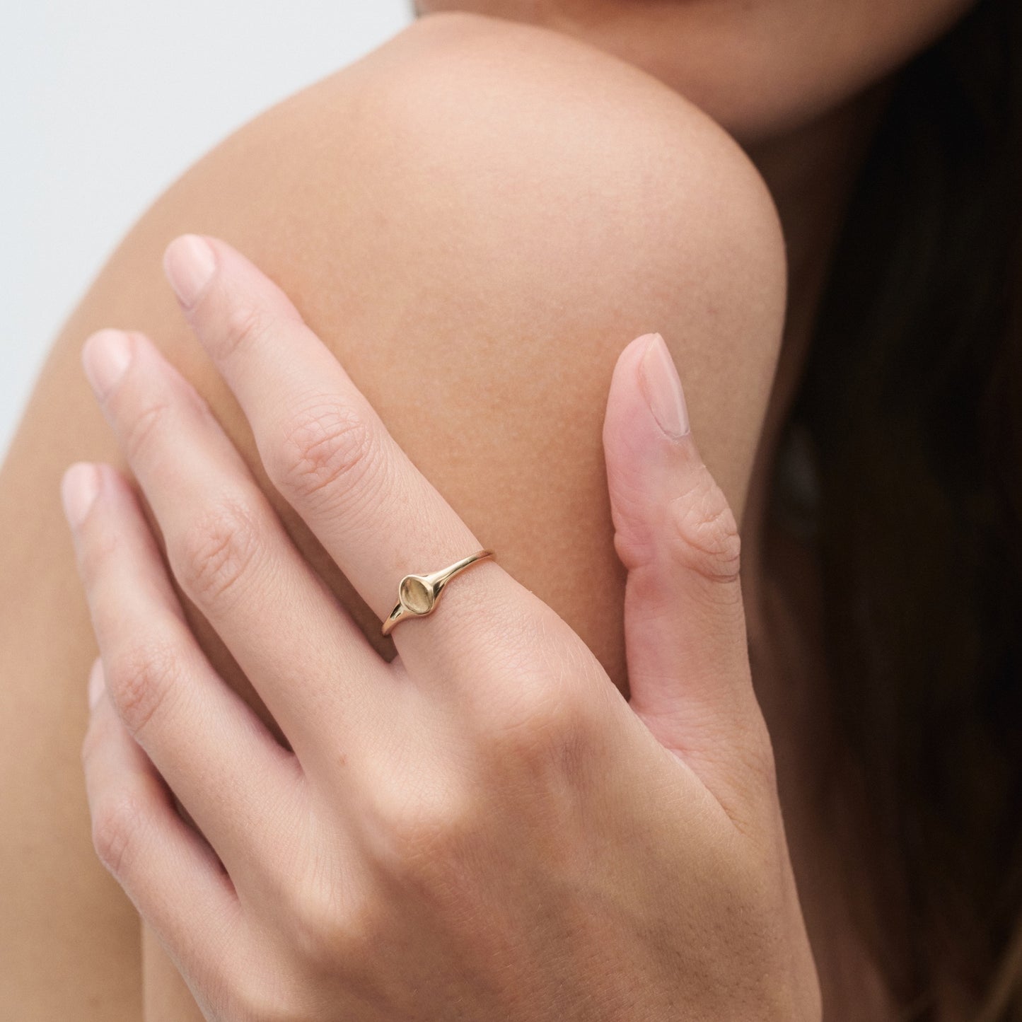 Hand wearing an elegant, thin gold ring with a unique heart design, gently touching a bare shoulder, showcasing luxury jewellery.