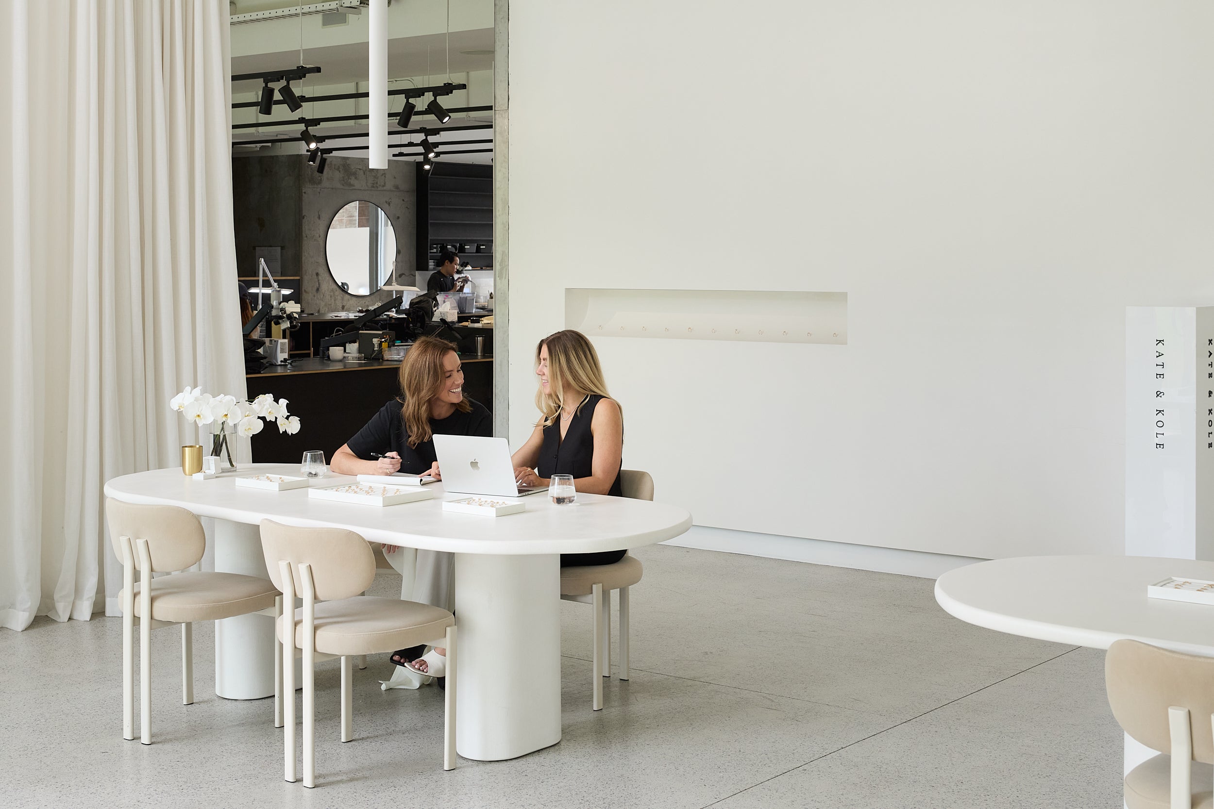 Kate & Kole founders, Sara and Maddy, sitting at a modern white dining table in a stylish interior setting.