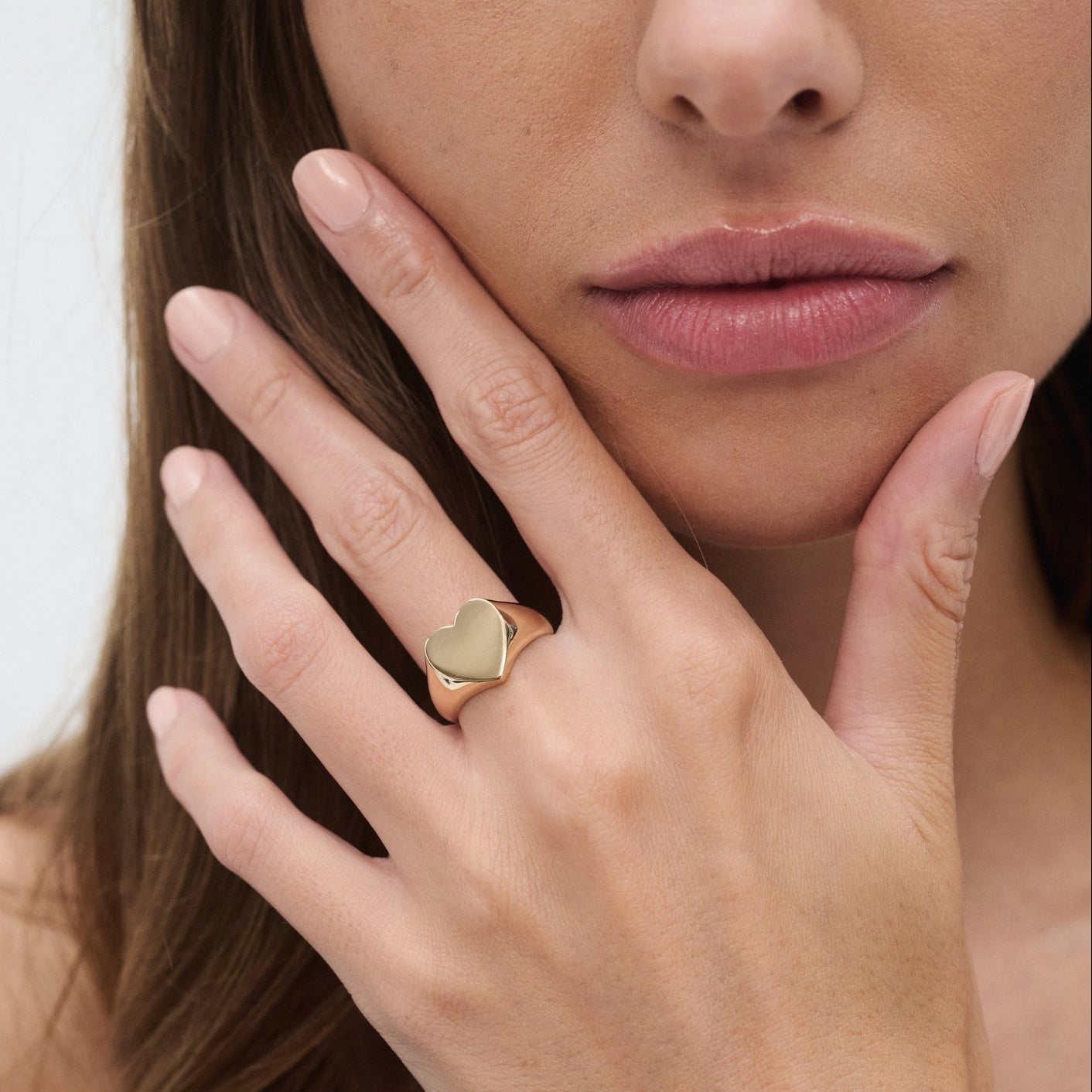 Woman's hand with a gold heart-shaped ring on her finger, touching her face. Luxury jewellery, elegance, minimalist style.
