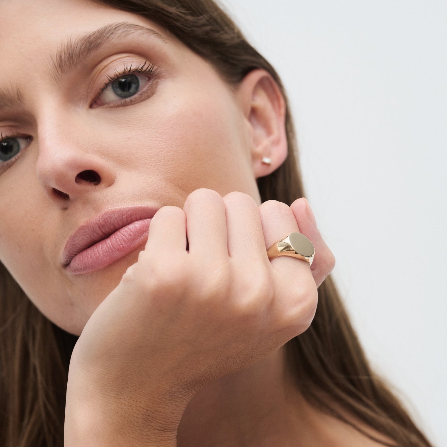 Woman showcasing luxury gold signet ring and delicate stud earring, set against a minimalist background.