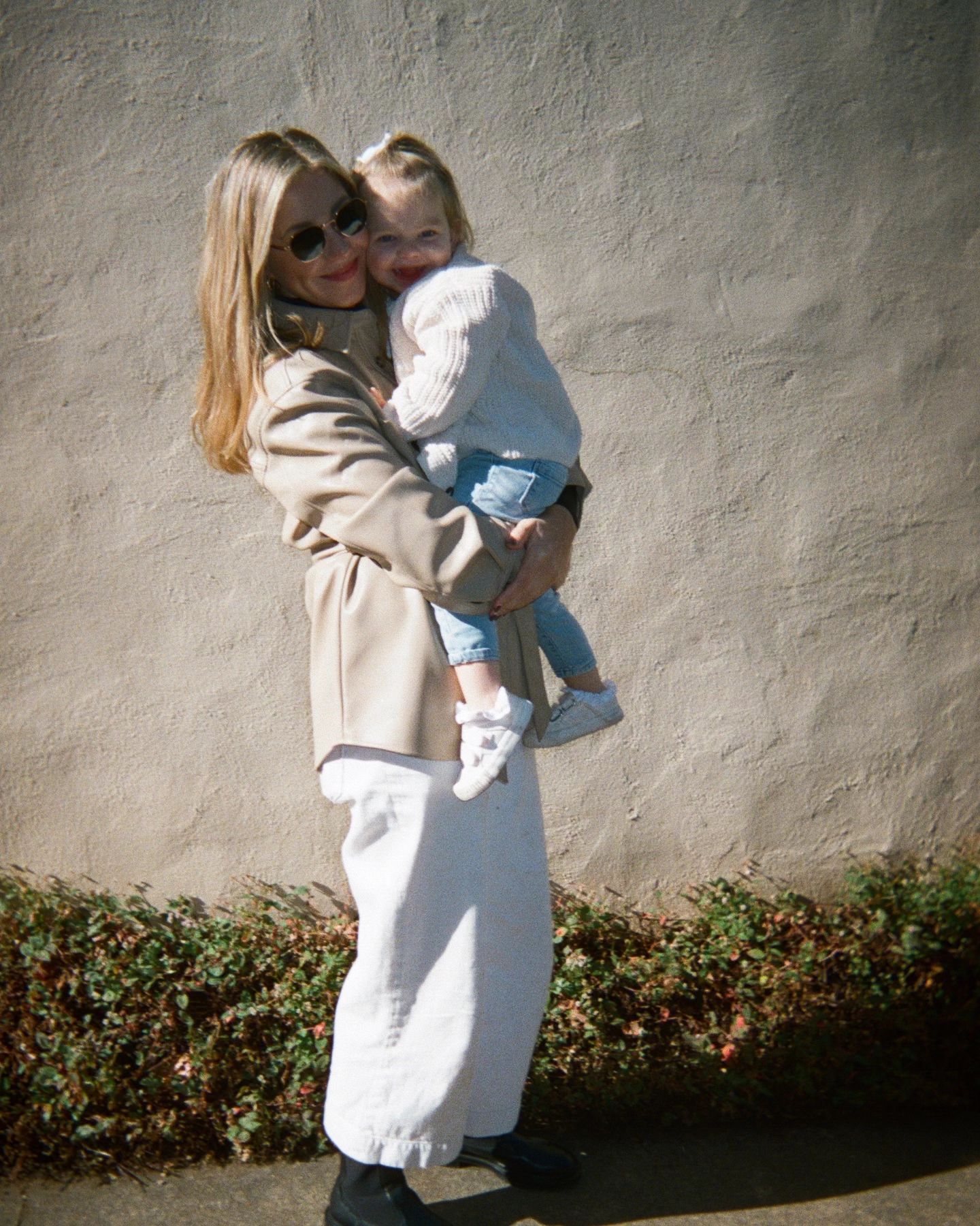 Mother in sunglasses and beige coat holding child in knit jumper against textured wall, standing on a path with plants, under bright sunlight.
