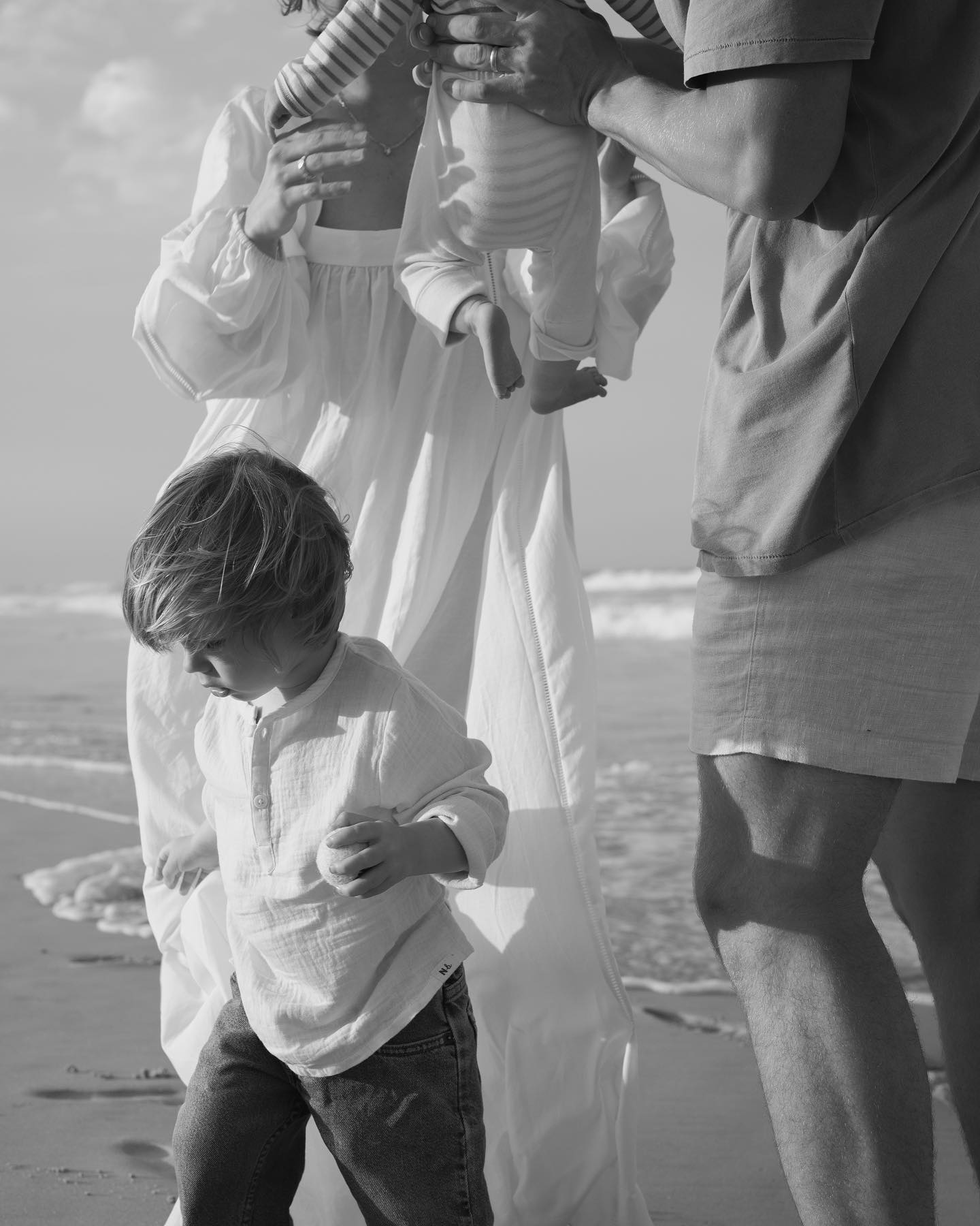 Family enjoying a beach day; child in foreground, adults and baby in flowy attire. Soft, monochrome tones create a serene, intimate atmosphere.