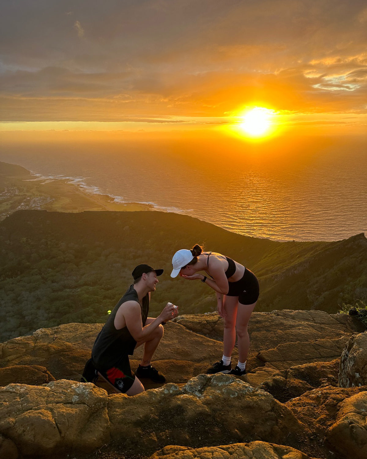 A couple at a mountain peak during sunset; one kneels, proposing with a ring. Ocean and coastal views fill the background.