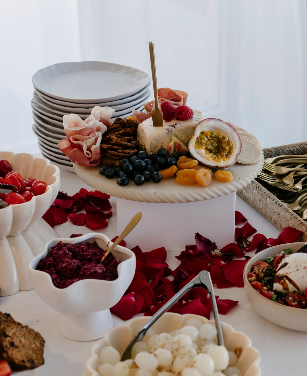 Elegant cheese platter with fruits, prosciutto, and crackers, surrounded by vibrant red rose petals on a white tablecloth.