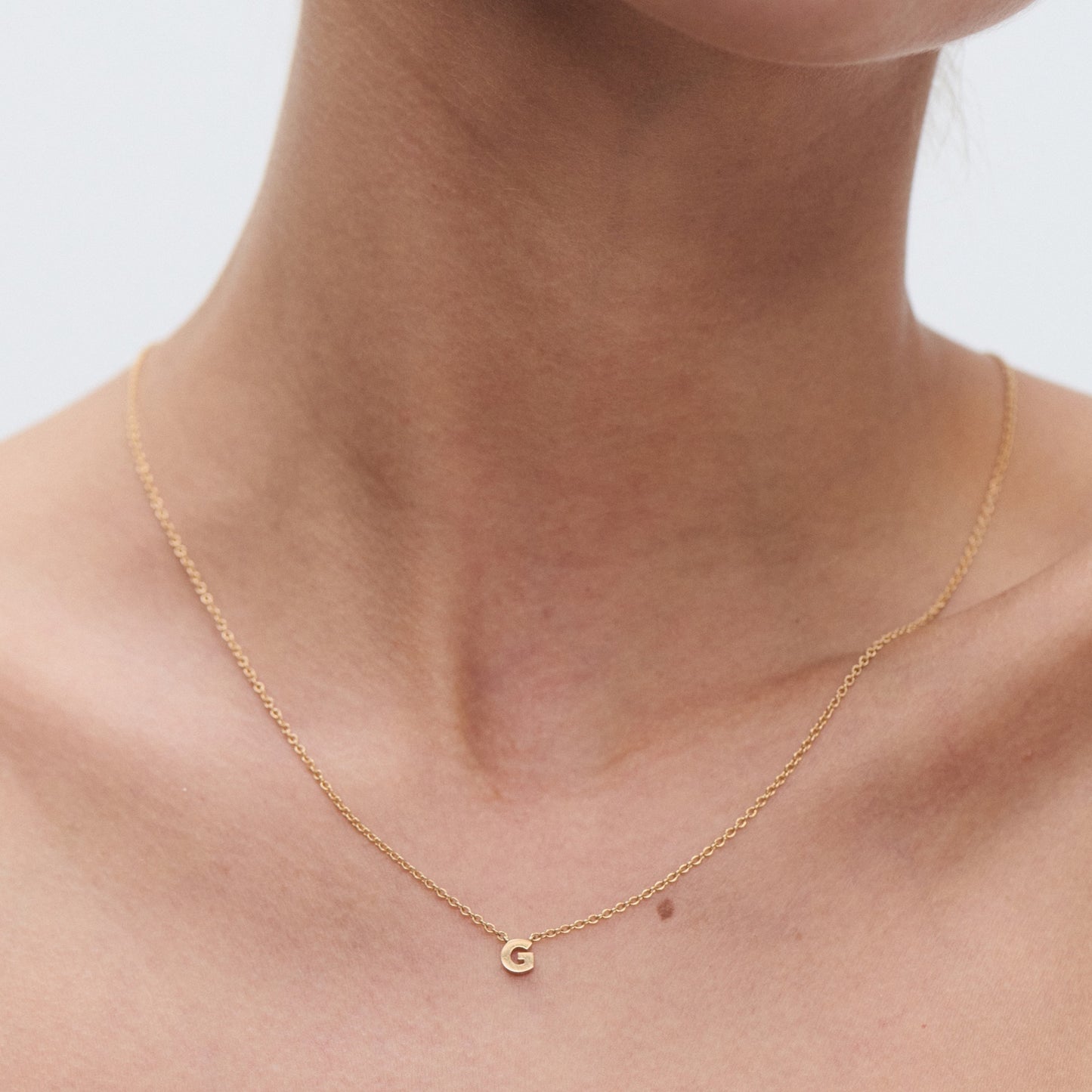 Delicate gold necklace with a small "G" pendant on a person's neck. Simple and elegant jewellery style.
