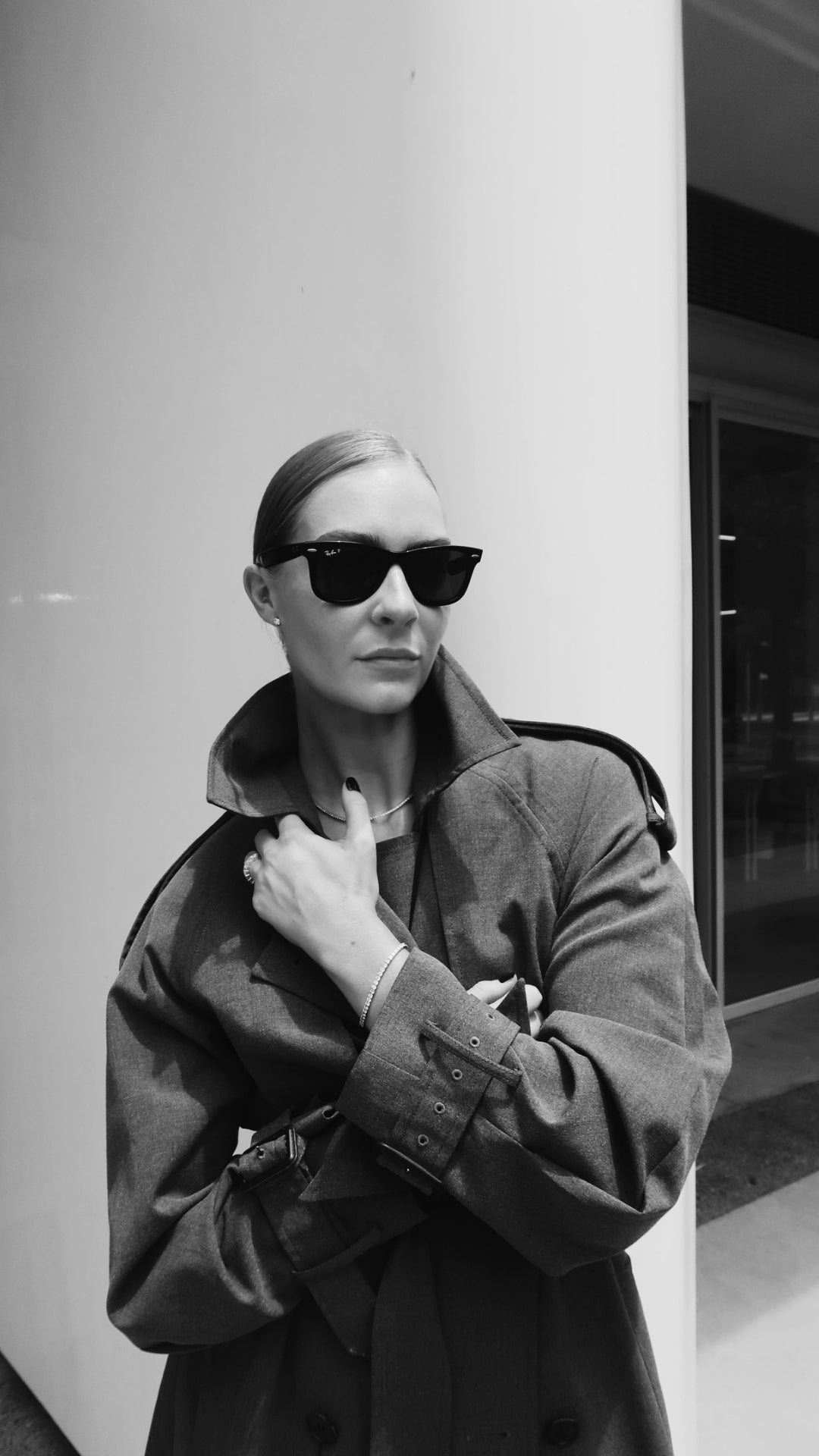 Stylish woman in sunglasses and trench coat, showcasing luxury jewellery with a sleek bracelet, posing confidently in an urban setting.