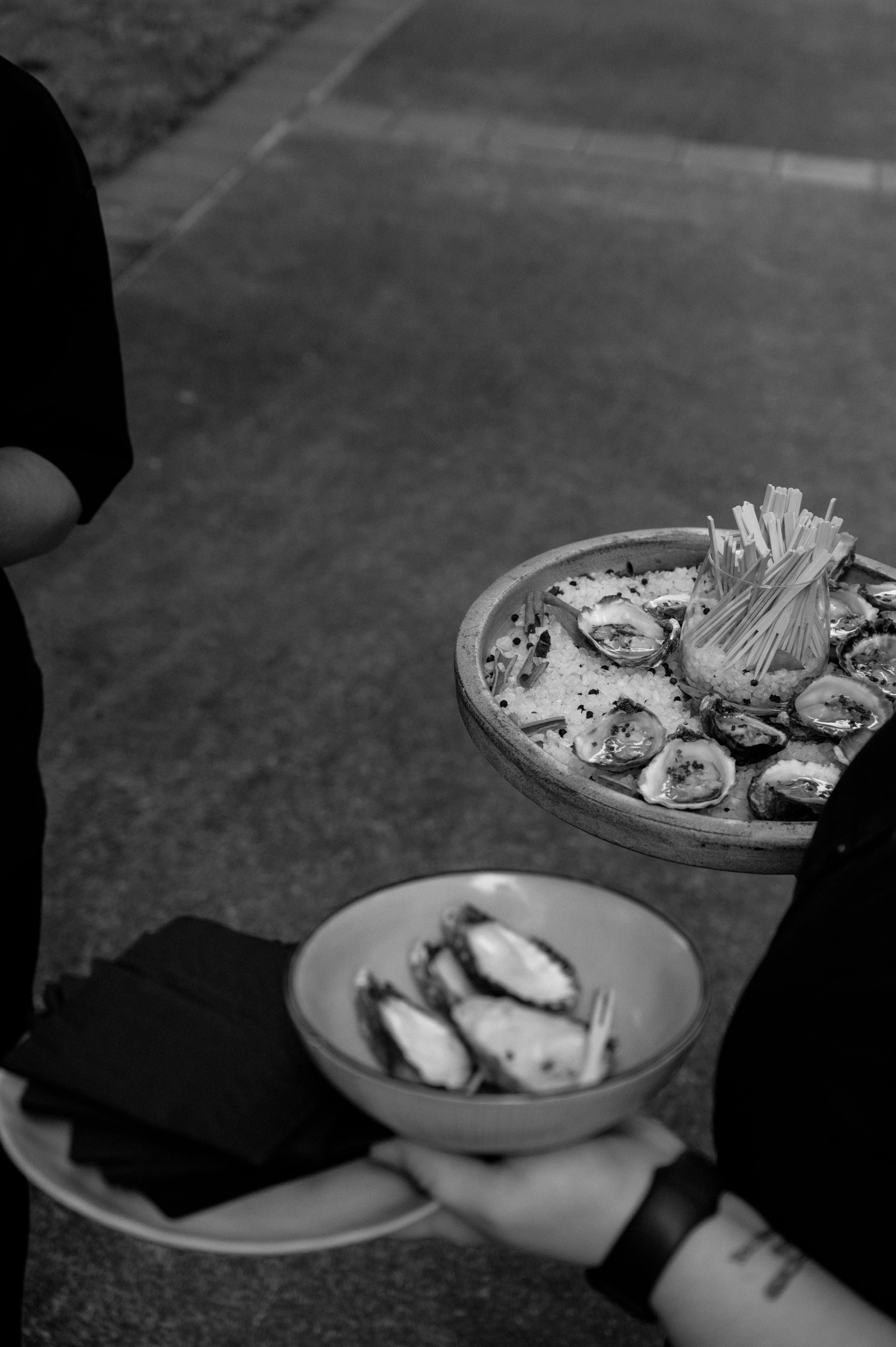 Black and white photo of gourmet oysters on a platter with toothpicks, held by a server, accompanied by a bowl of bread and napkins.