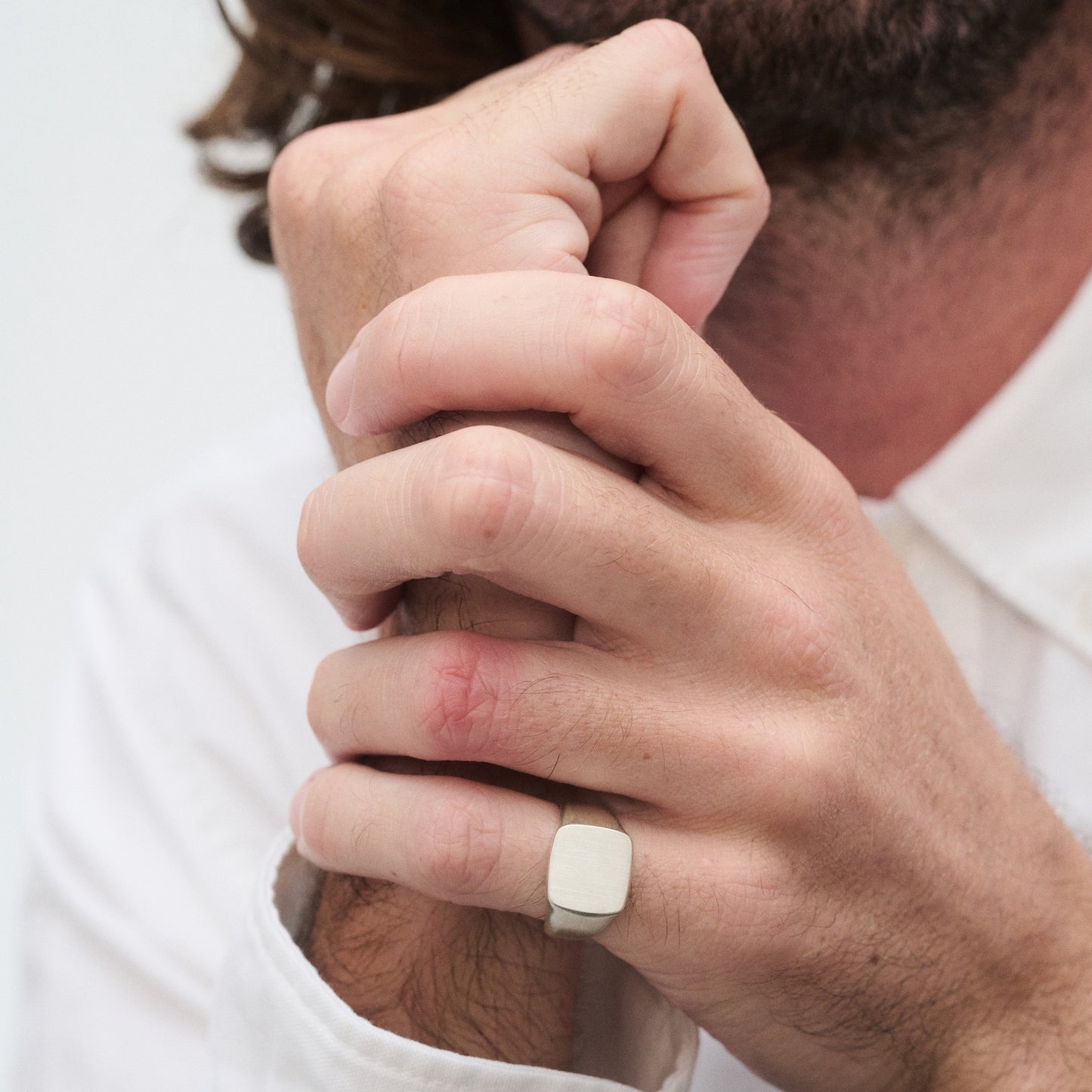 Man in a white shirt wearing a minimalist silver signet ring on his finger, arm rested against his chin, showcasing luxury jewellery style.