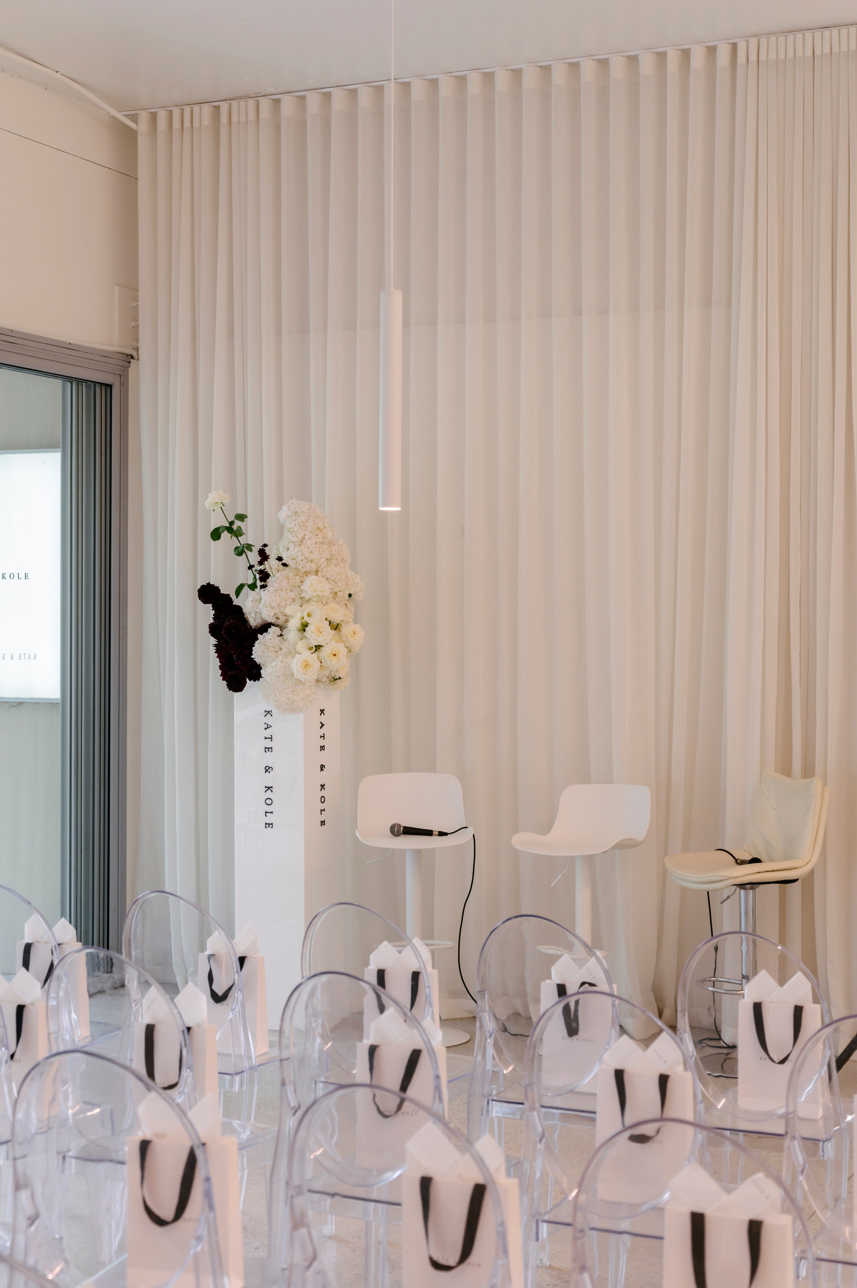 Elegant event setup with clear chairs, white floral arrangements, and podium. Minimalist design, luxury vibes, likely a jewellery launch.