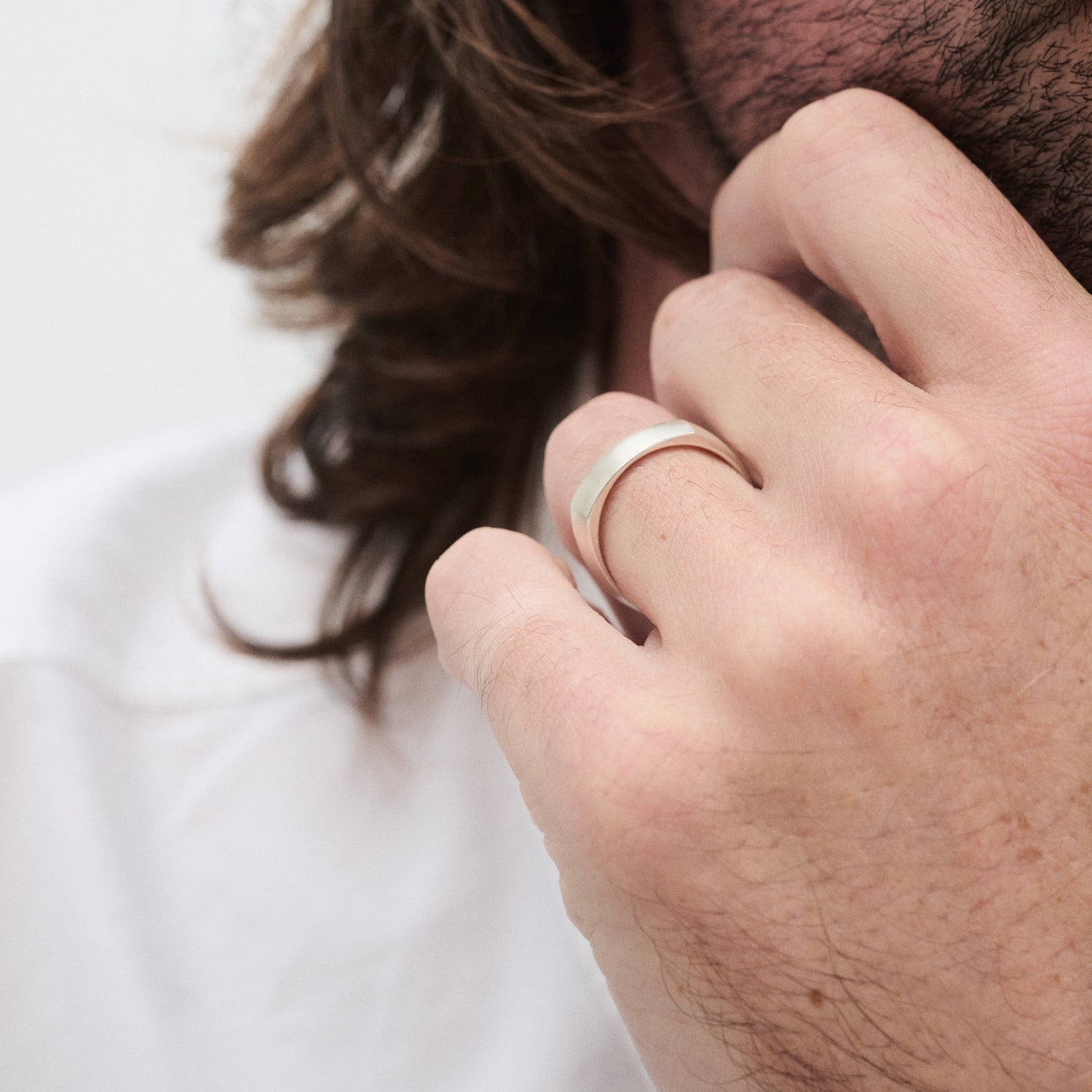 Man with long hair wearing a silver band ring on his finger, touching his chin thoughtfully. White shirt in background.