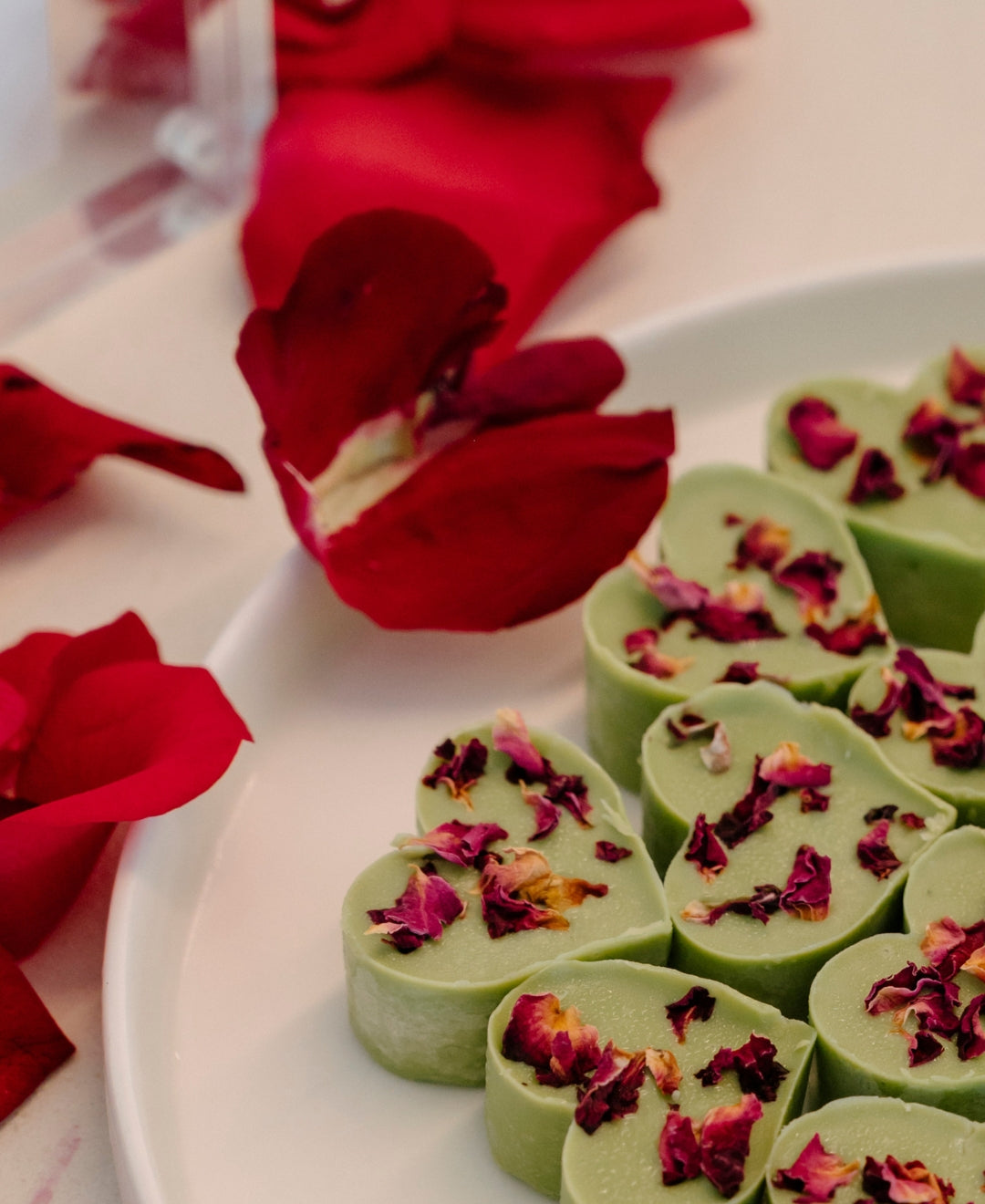 Heart-shaped green chocolates adorned with dried rose petals on a plate, surrounded by vibrant red rose petals. Luxurious and elegant presentation.