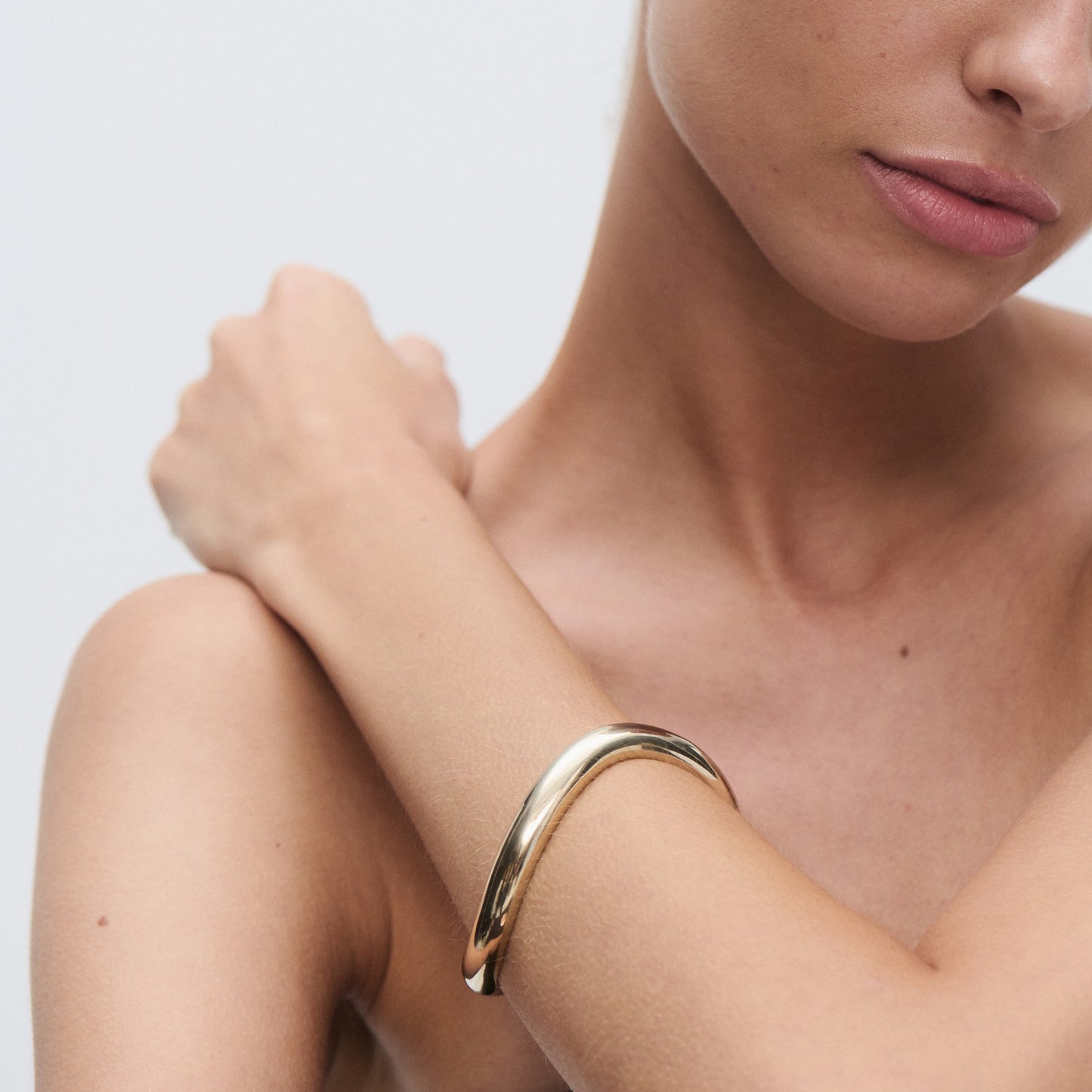 Woman's shoulder and arm with a sleek, polished gold bangle bracelet, showcasing minimalist luxury jewellery. Light background.[text overlay: Bold] [text colour: #000000]
