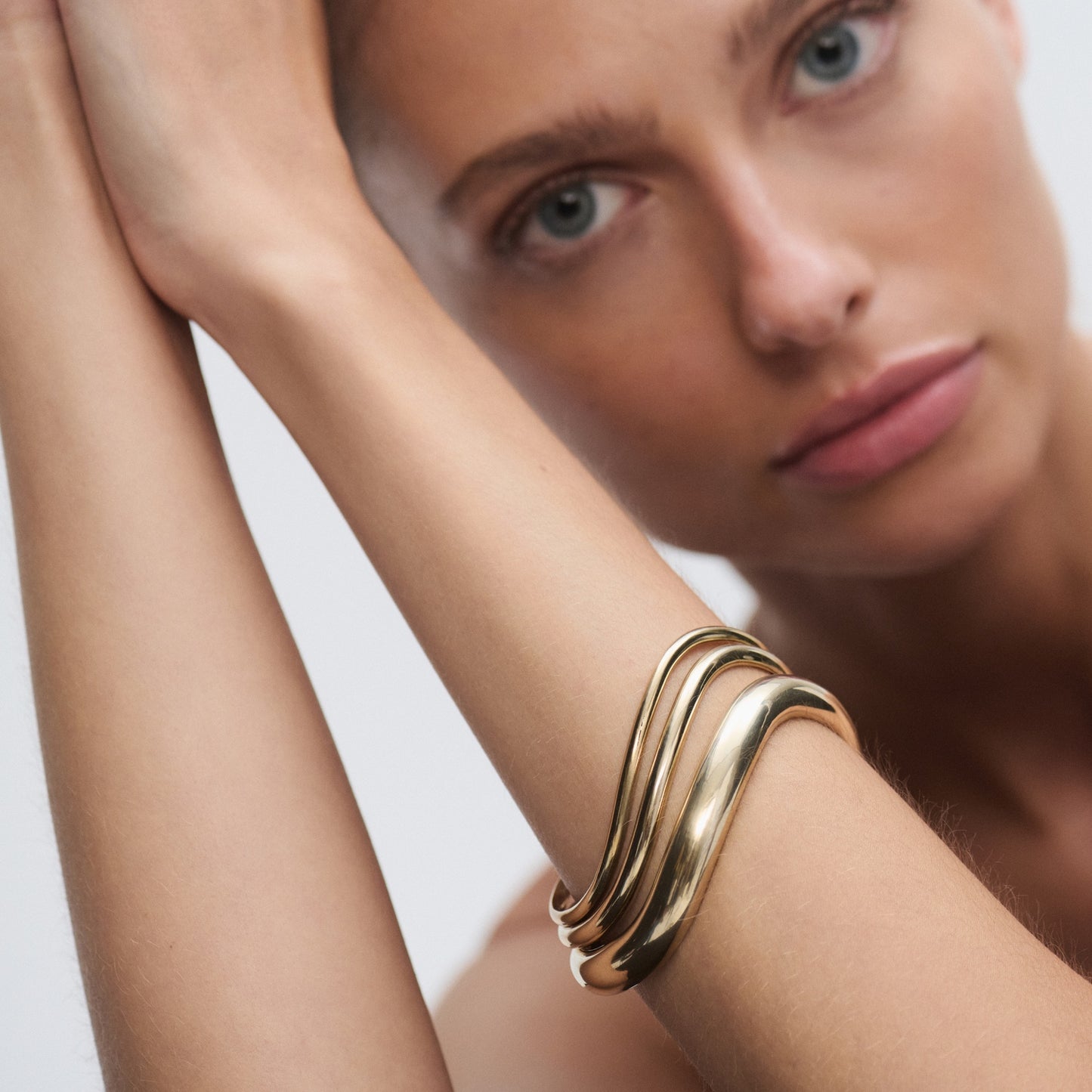 Model wearing an elegant, wavy gold bracelet; luxury jewellery close-up. Pale blue eyes, focused expression, soft background. [text overlay:] [text colour: #000000]
