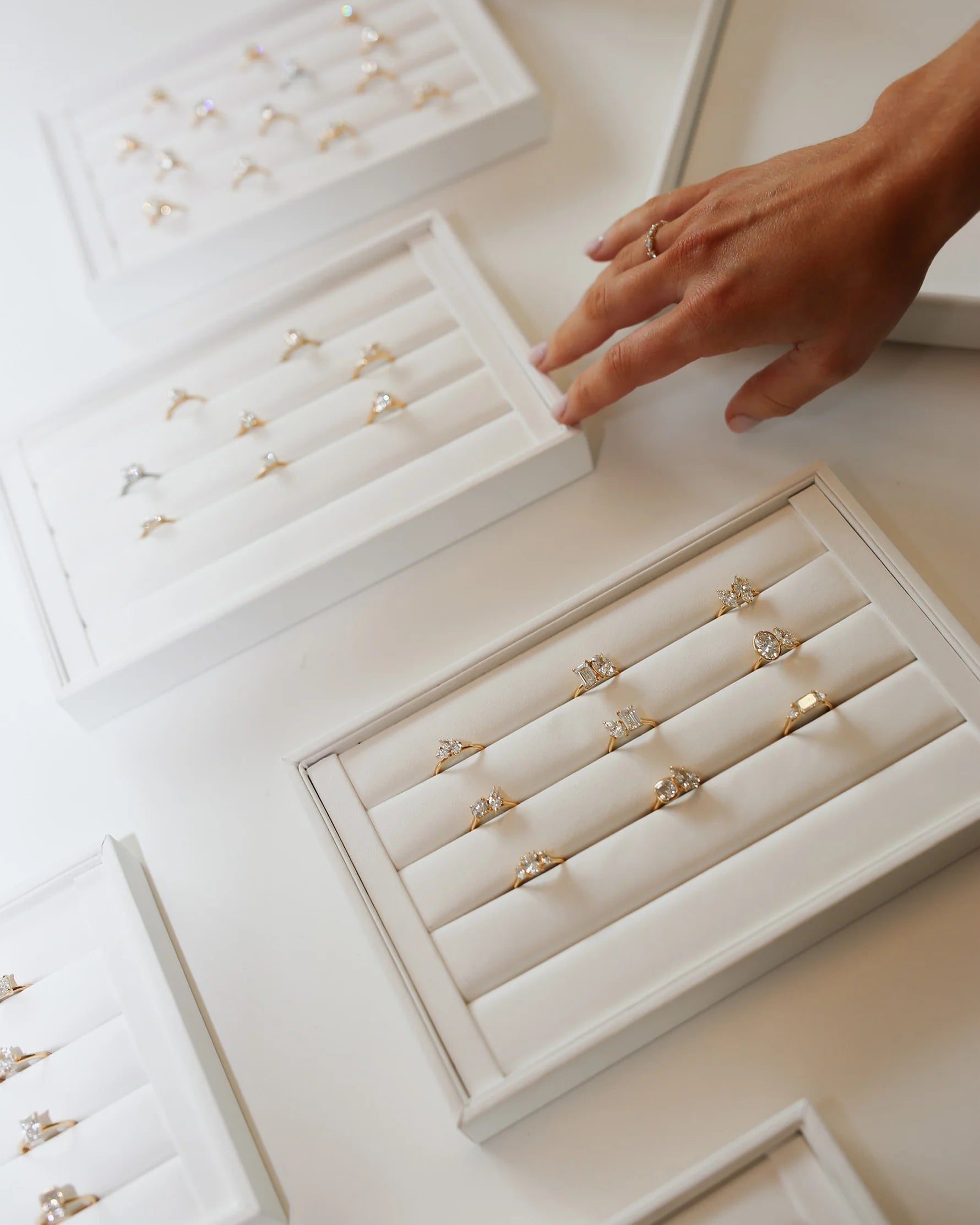 Hand selecting elegant gold and diamond rings from white jewellery trays, showcasing luxury engagement rings on a white surface.