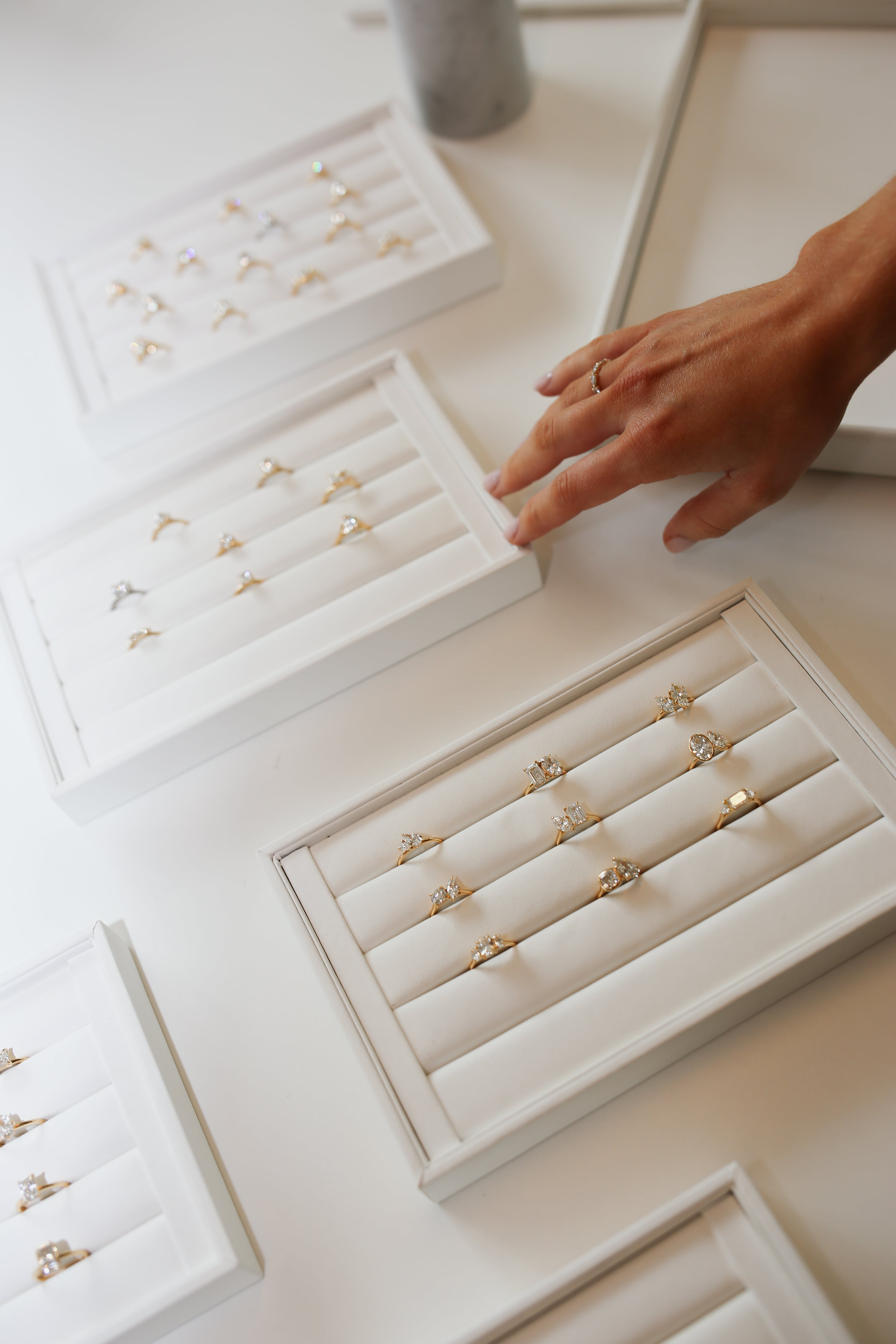 Luxury diamond rings displayed in elegant white trays on a table; a hand reaches for a ring.