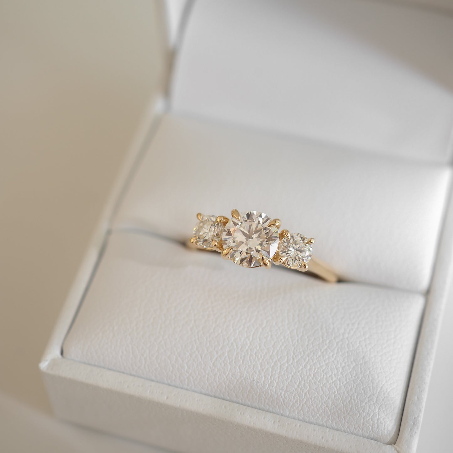 A gold engagement ring with a large round diamond flanked by two smaller square diamonds is displayed in a white jewellery box.