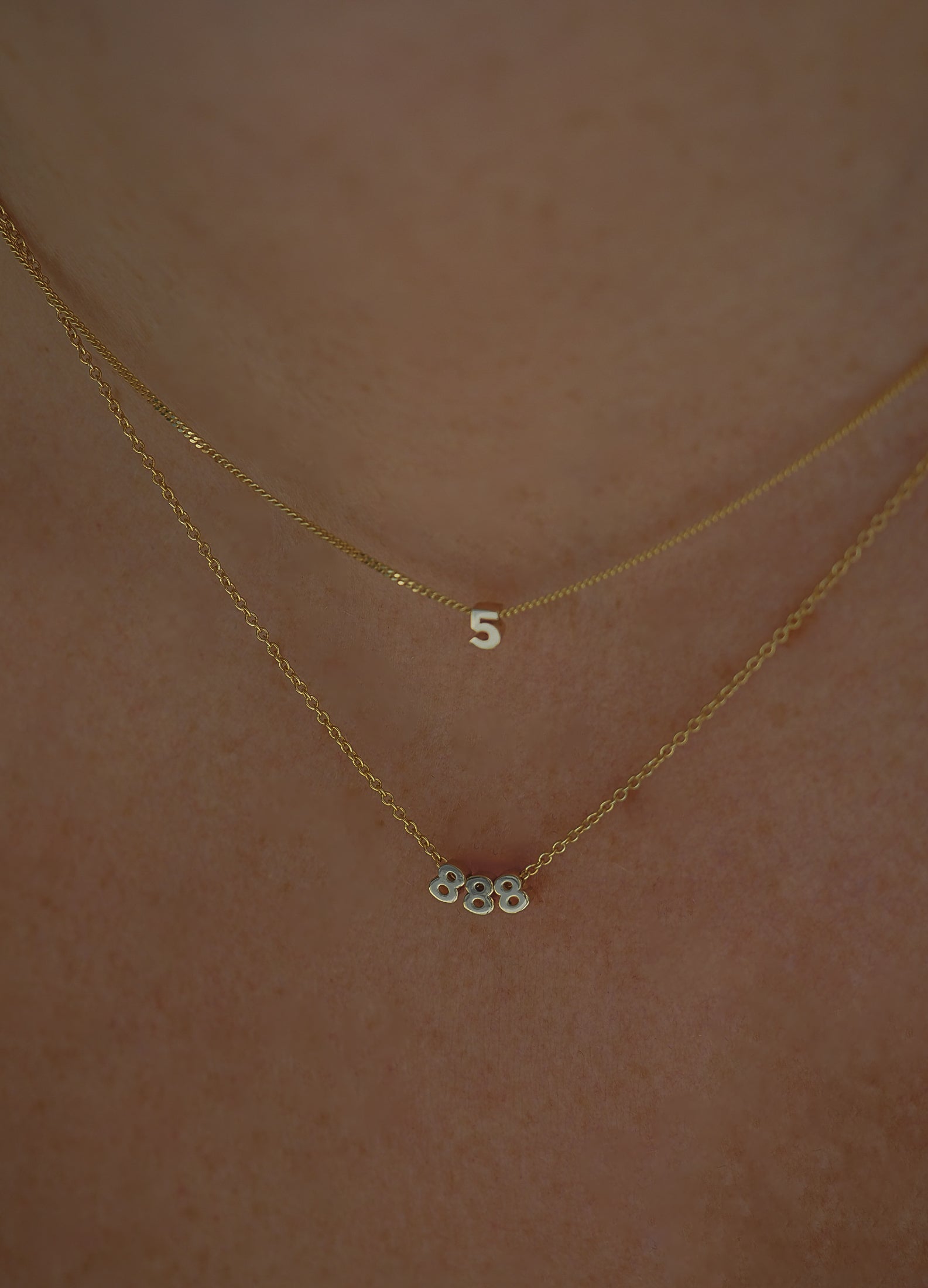 Two gold necklaces with numerical pendants "5" and "888" on a person’s neck, highlighting luxury jewellery design.