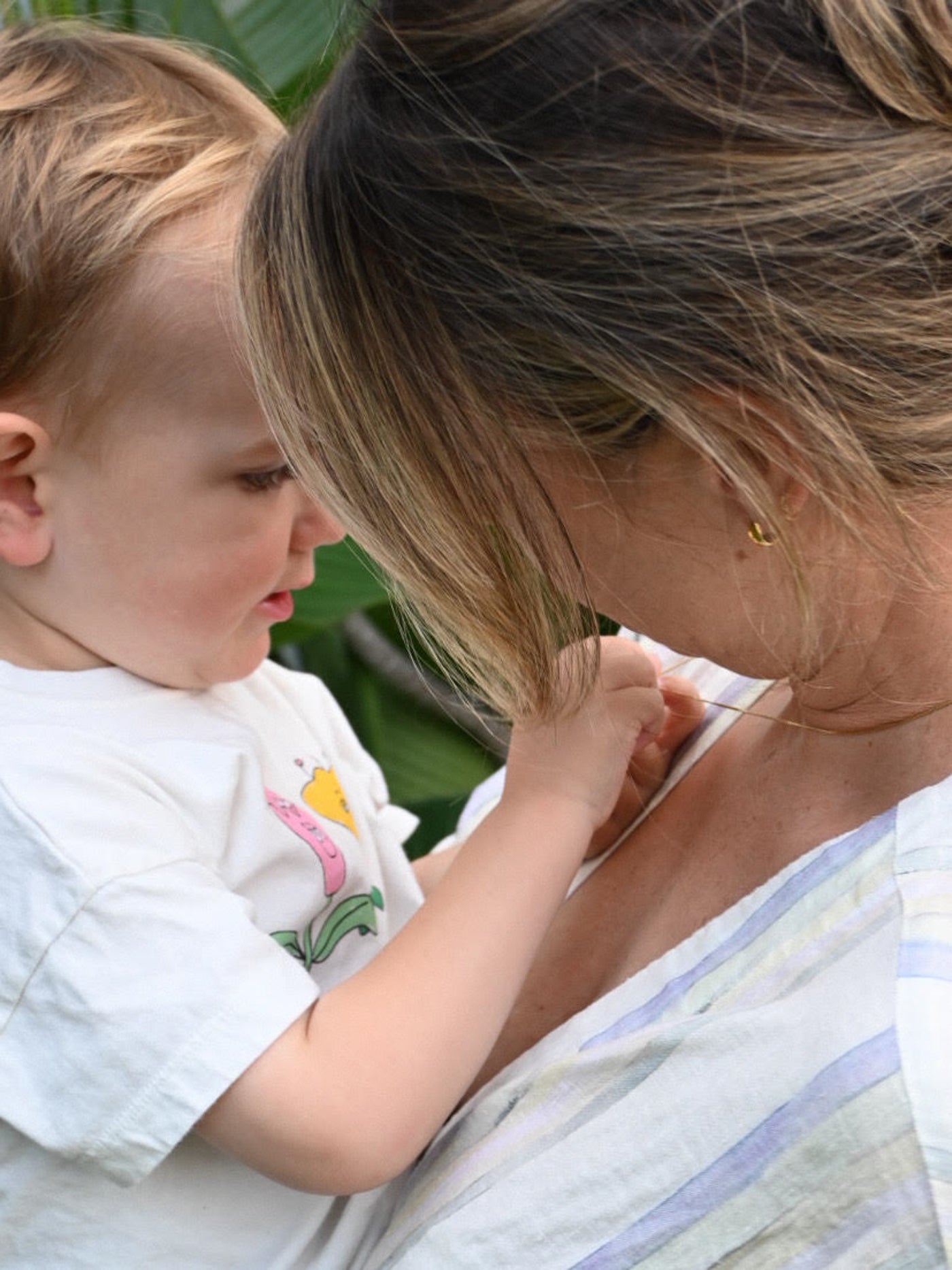 A toddler in a white shirt looks closely at a woman's jewellery while she wears a striped top, highlighting a personal and intimate moment.