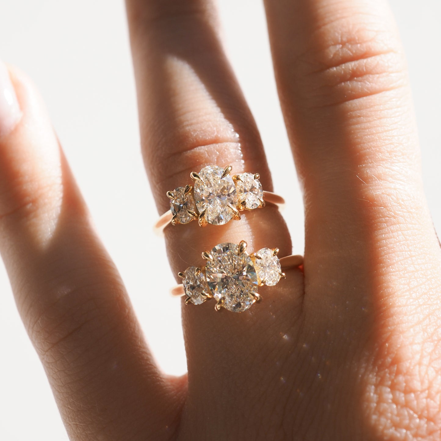 Hand wearing two gold rings with large, sparkling diamonds in sunlight. High-end luxury jewellery showcasing elegance and sophistication. [text overlay: 1ct Centre Stone (Top) 1.5ct Centre Stone (Bottom) ] 
