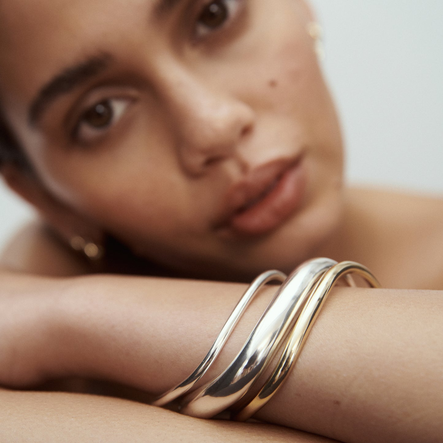 Close-up of a woman resting her head on her arm, showcasing sleek, interwoven silver and gold high-end bangles, adding a touch of luxury. [text overlay: Fine (Left), Bold (Middle) and Medium ( Right)] 