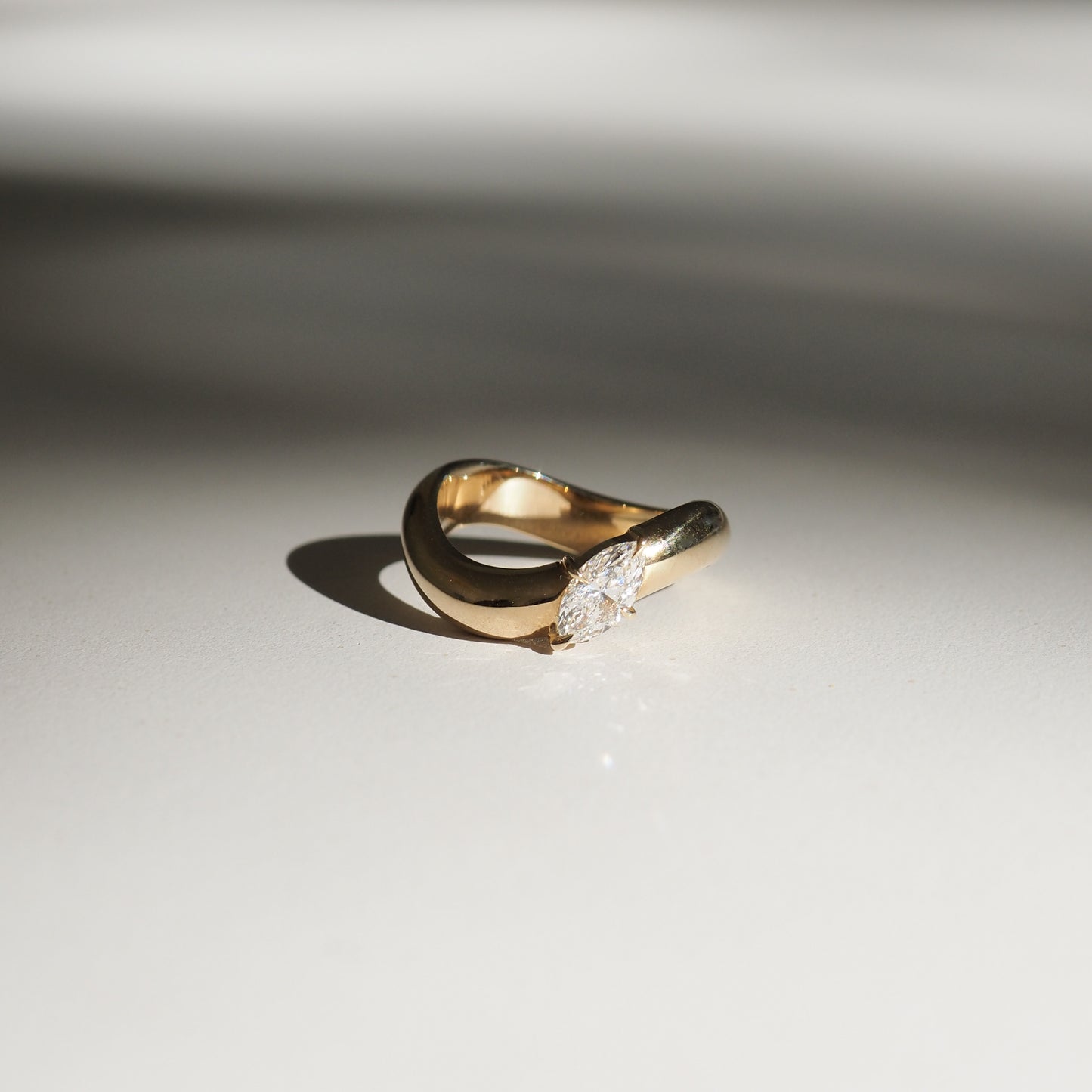 Gold ring with a marquise diamond set in a unique twist design, casting a soft shadow on a neutral surface. Luxurious and elegant jewellery.