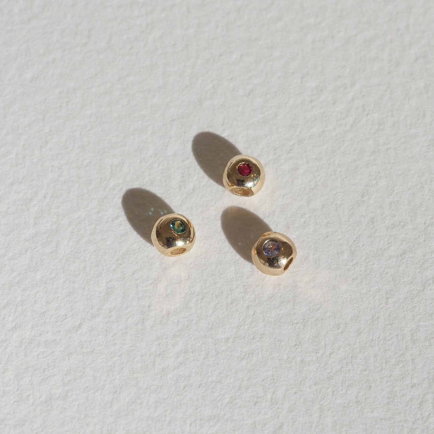 Three small gold stud earrings with red, green, and grey gemstones on a textured white surface, casting soft shadows.[text overlay: Emerald (left), Ruby (middle and Tanzanite (right)] 
