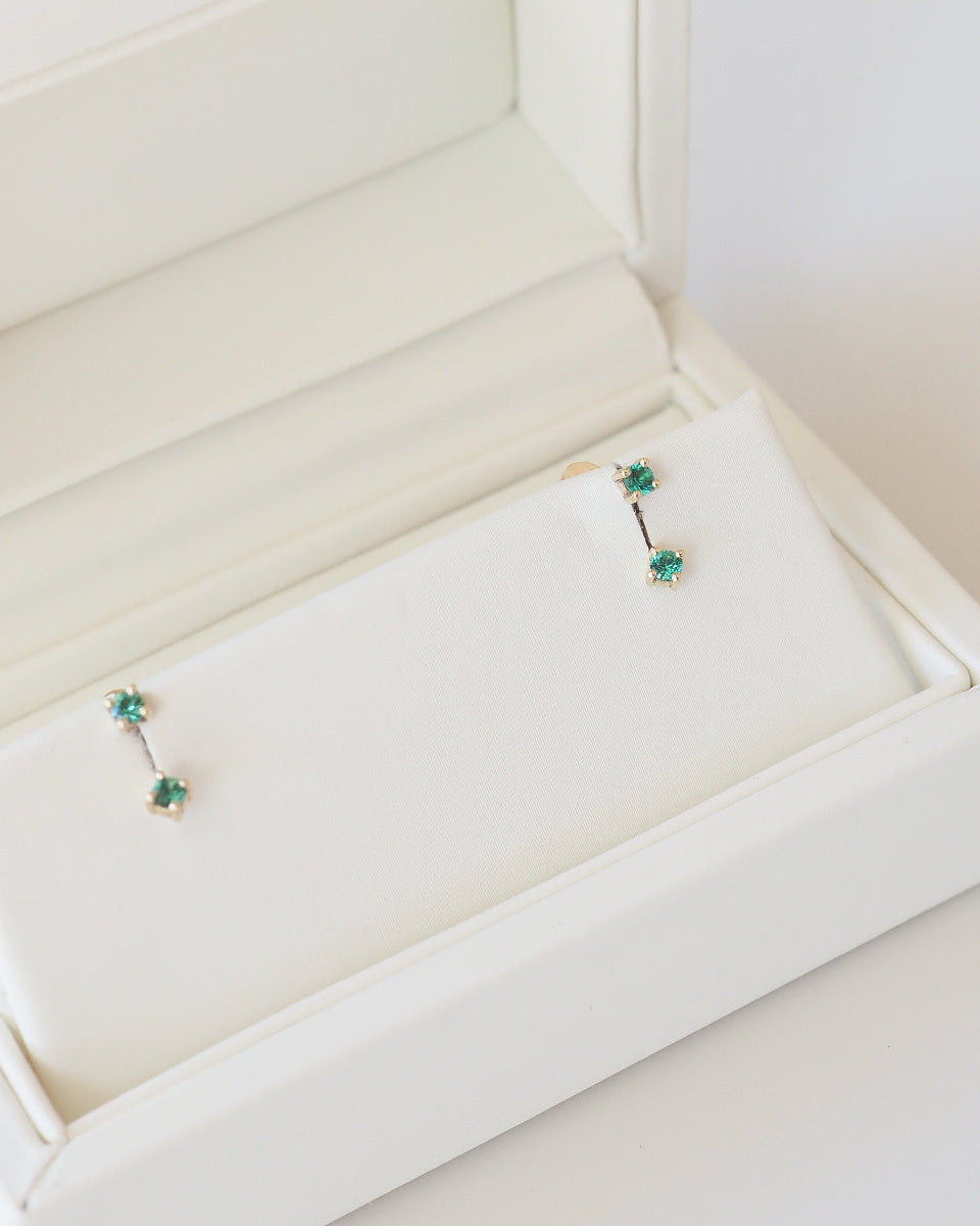 Pair of earrings with green gemstones in a white jewelry box [text overlay: Tiny Birthstone Jackets and Studs]