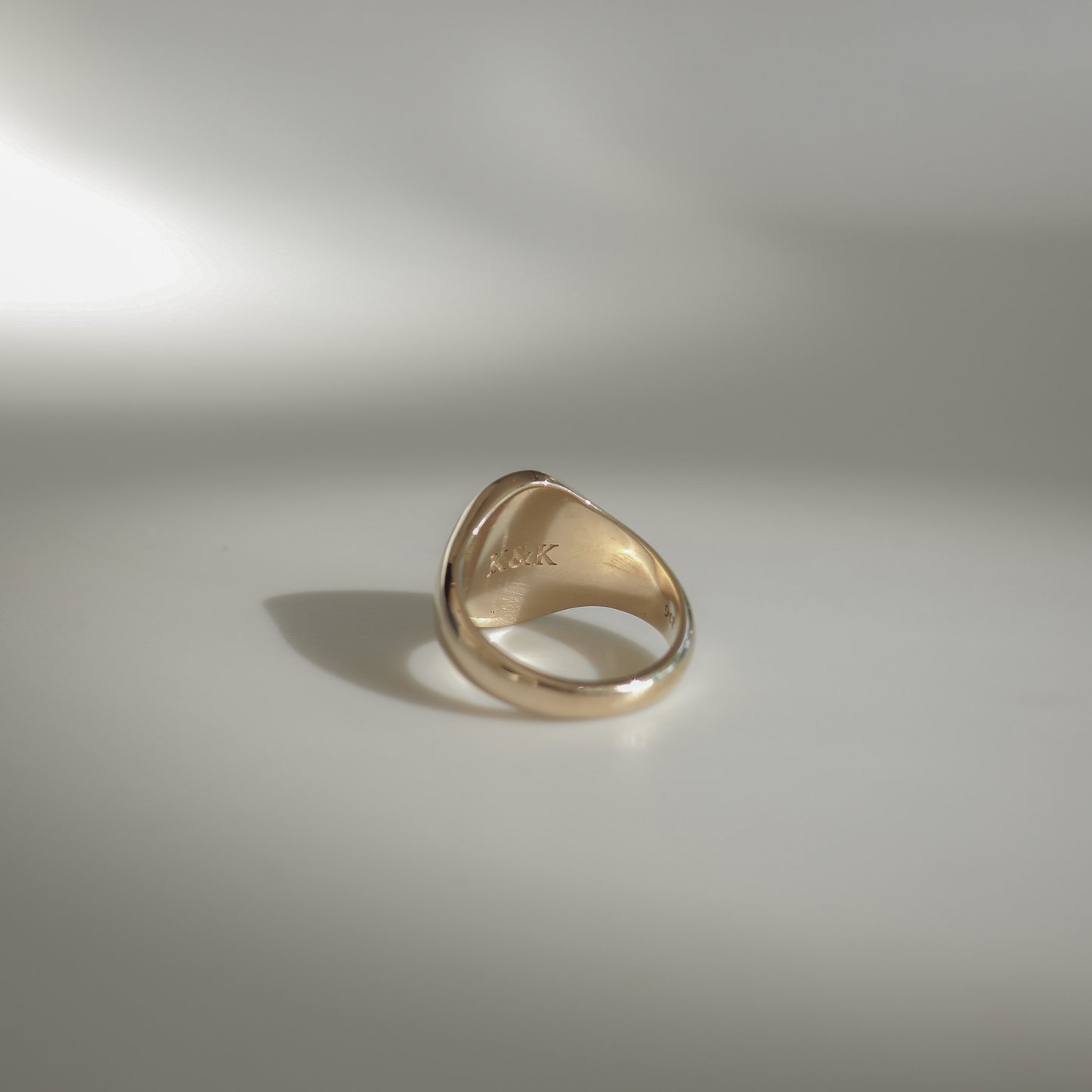 Gold ring with engraved initials "A&K" on a smooth surface, softly lit against a neutral background. Elegant and refined luxury jewellery.