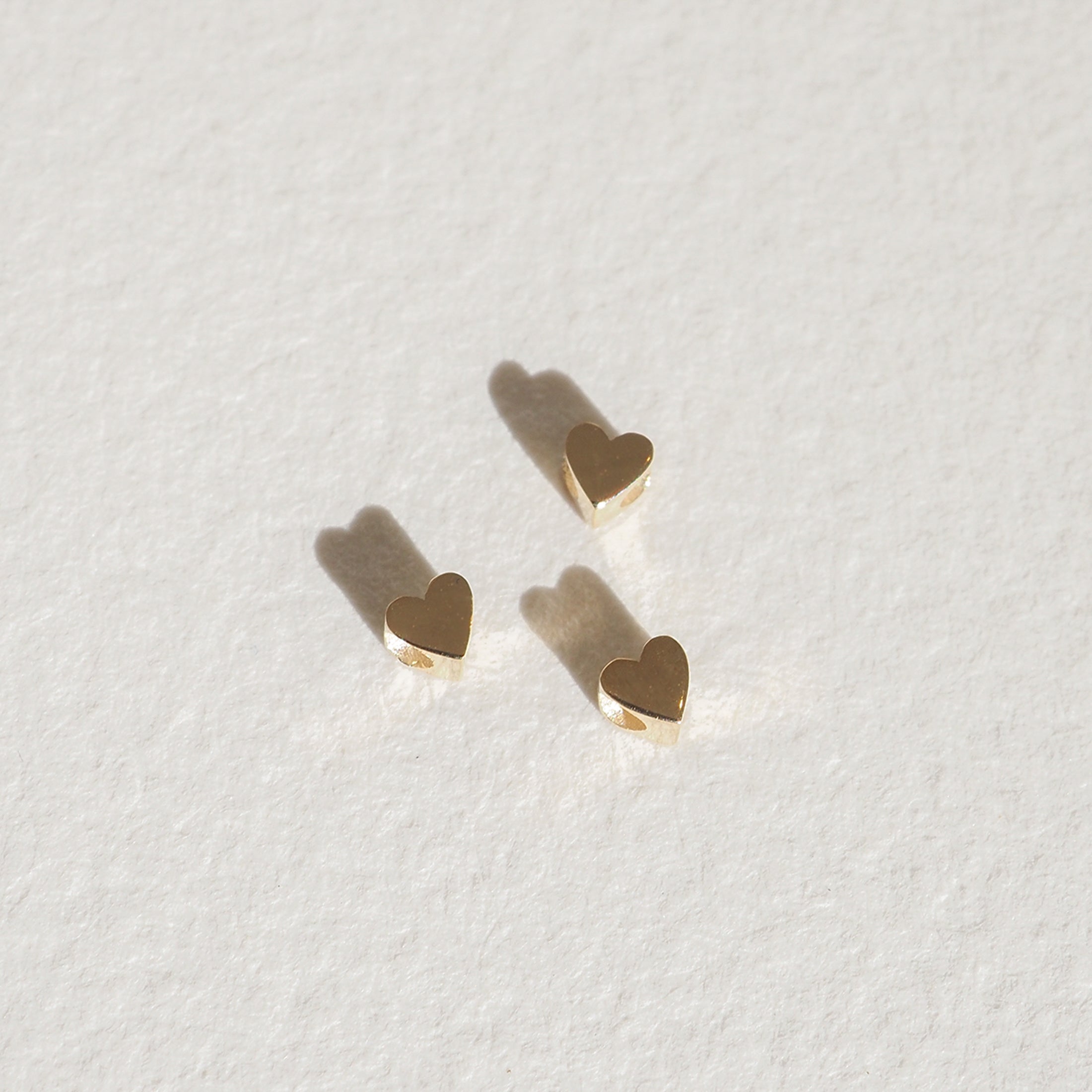 Three small gold heart-shaped earrings on a textured, light background casting soft shadows. Luxurious jewellery minimalism.