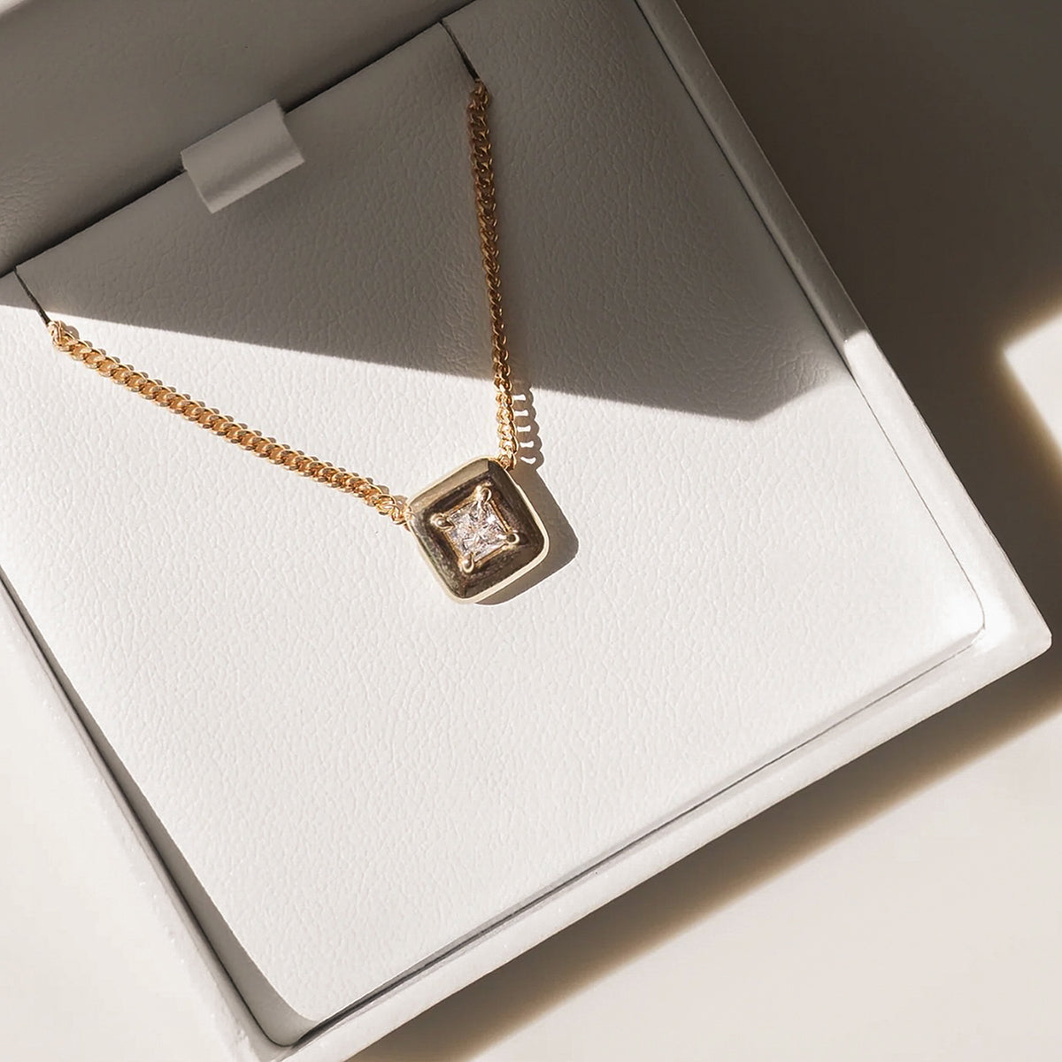 Gold necklace with a square diamond pendant in a white jewellery box, bathed in natural light. Luxurious, elegant design.